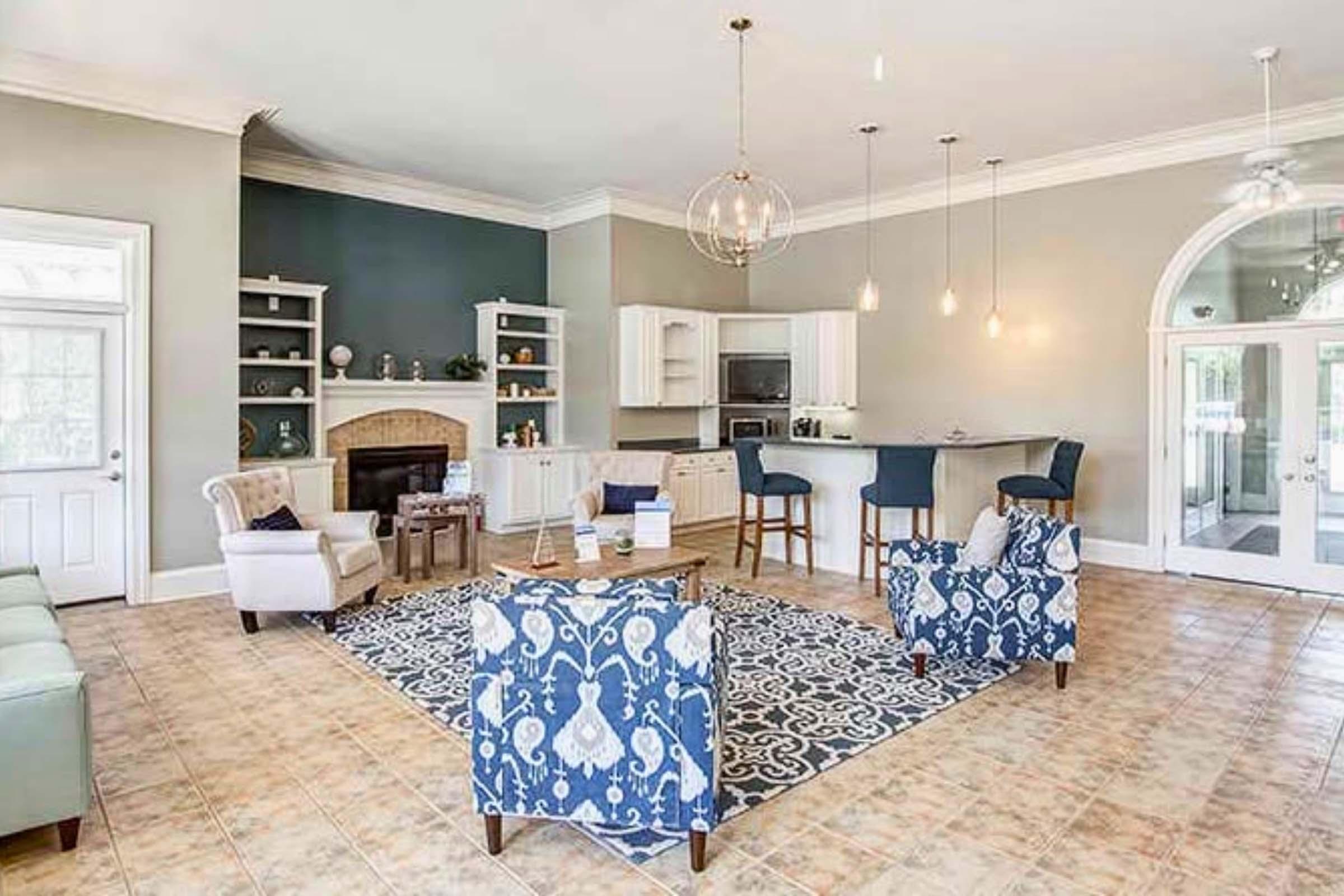 A spacious and modern living area featuring a cozy seating arrangement with patterned chairs and a light sofa. A neutral color scheme complements the room, which includes a fireplace and an open kitchen space with bar stools. Natural light floods in through large windows, creating a welcoming atmosphere.