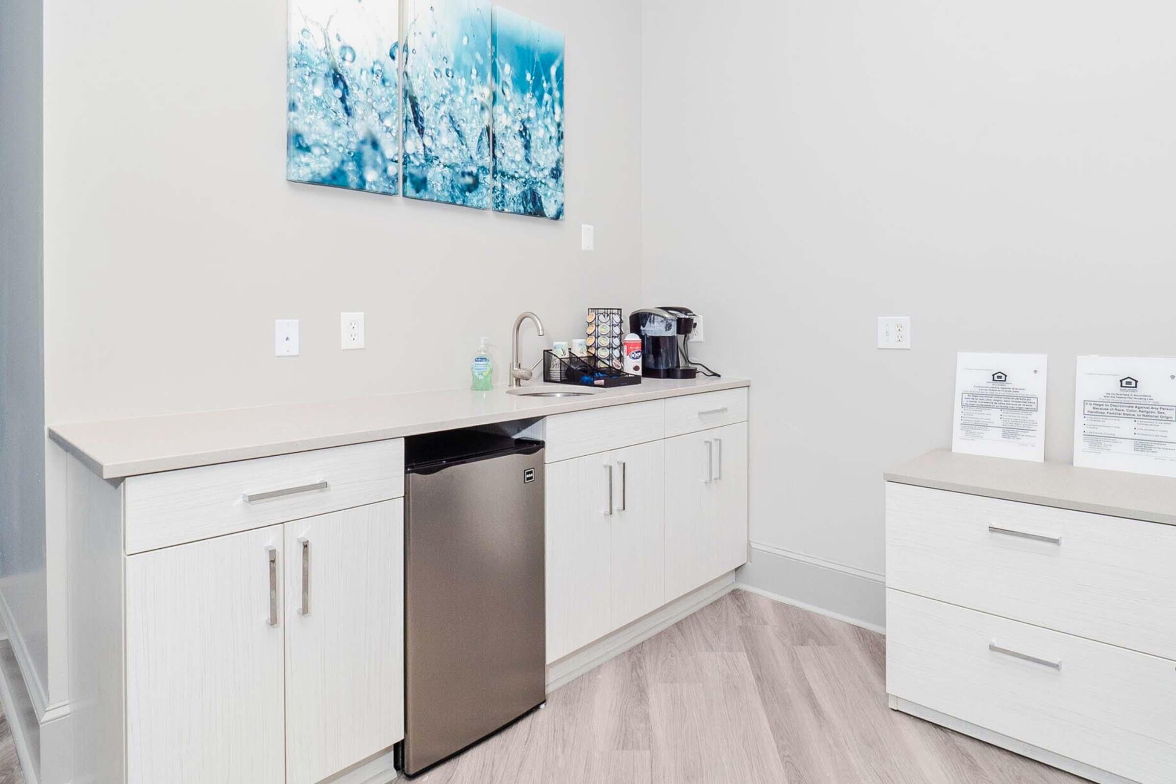 A modern kitchen space featuring light-colored cabinetry, a small stainless steel refrigerator, a coffee maker, and a countertop with a sink. Above the counter, there are two framed artwork pieces depicting water droplets. The flooring is light wood, enhancing the contemporary look.