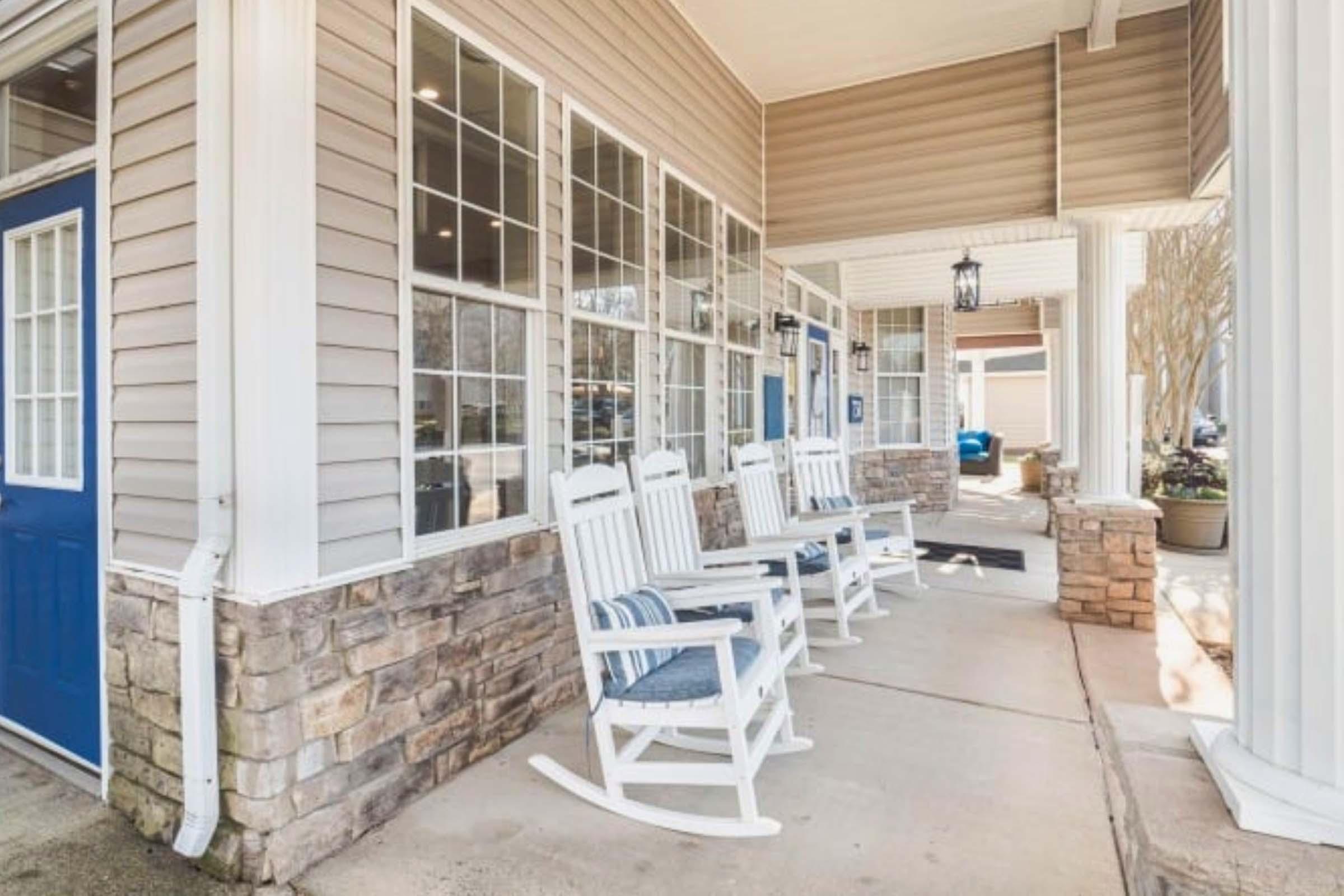 A porch area featuring several white rocking chairs aligned on a concrete surface next to large windows. The exterior wall is a mix of beige siding and stone. A blue door is visible, and there are potted plants adding a decorative touch to the setting. Soft natural light brightens the space.