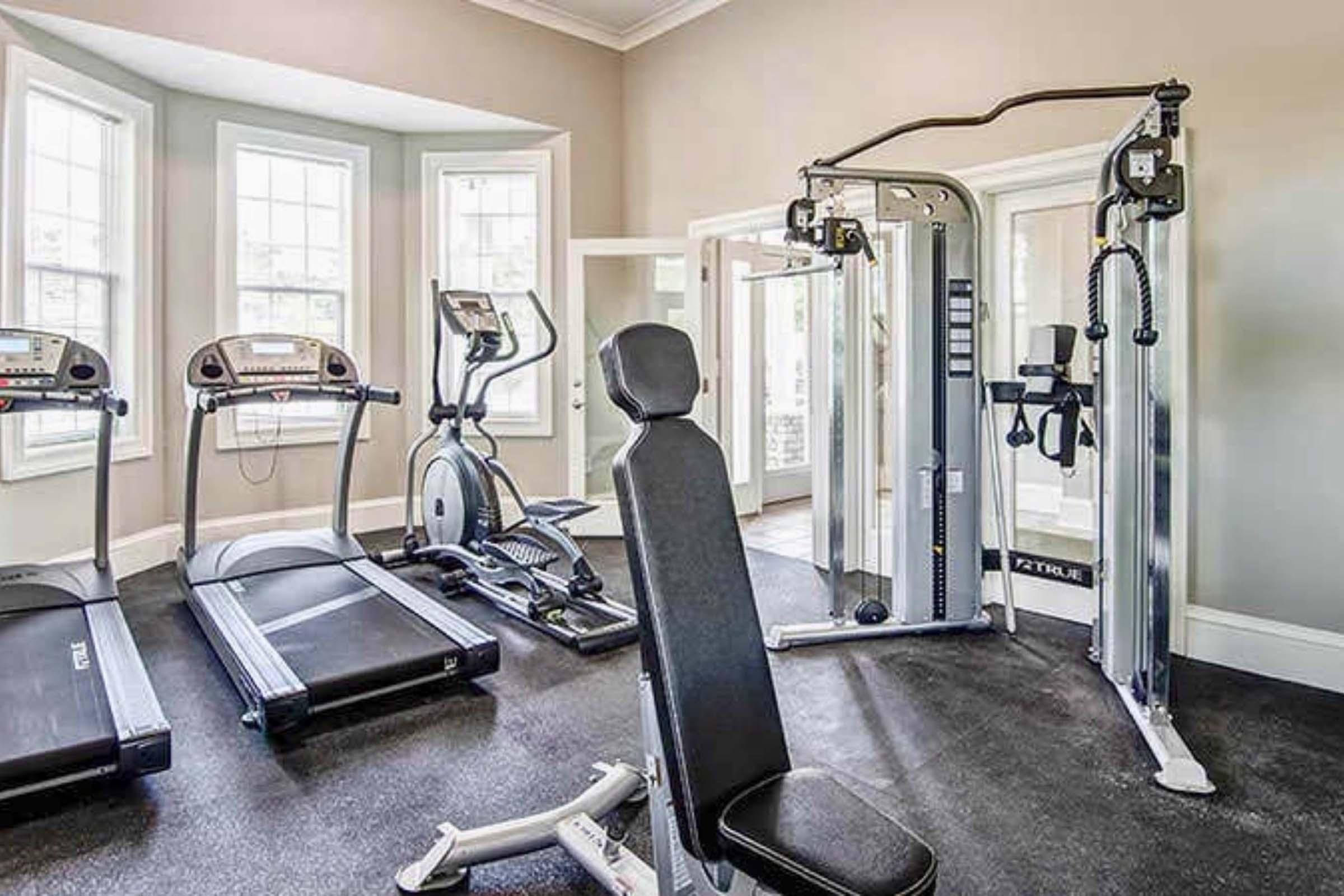 A modern home gym featuring two treadmills, an elliptical machine, and a multi-station weight machine. The space is well-lit with large windows, and the flooring is black rubber. A workout bench is positioned in the center, creating an inviting and functional workout area.