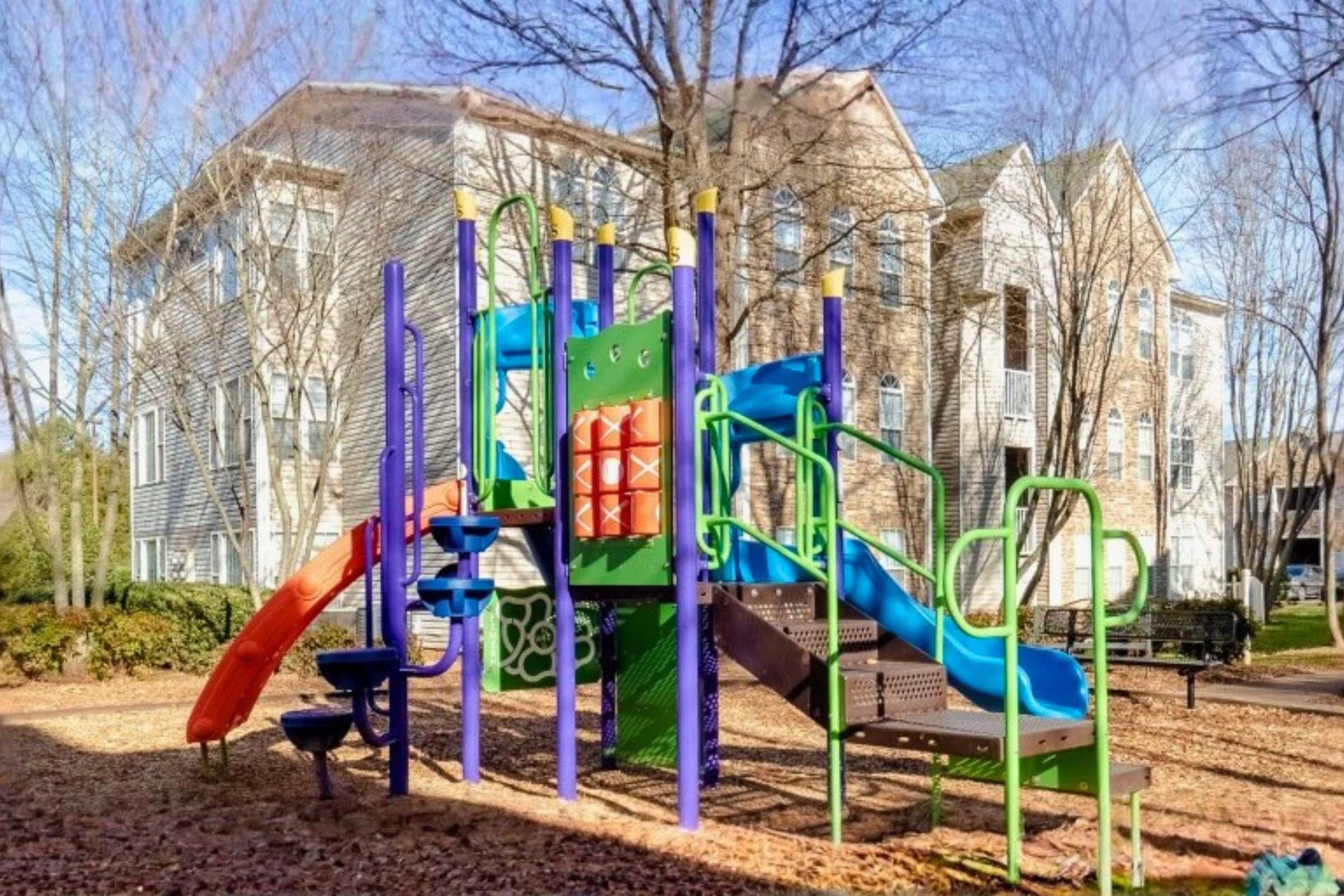 A colorful playground with slides, climbing structures, and a tunnel, surrounded by trees and residential buildings in the background. The playground features purple, green, and orange elements and is situated on a bed of wood chips.