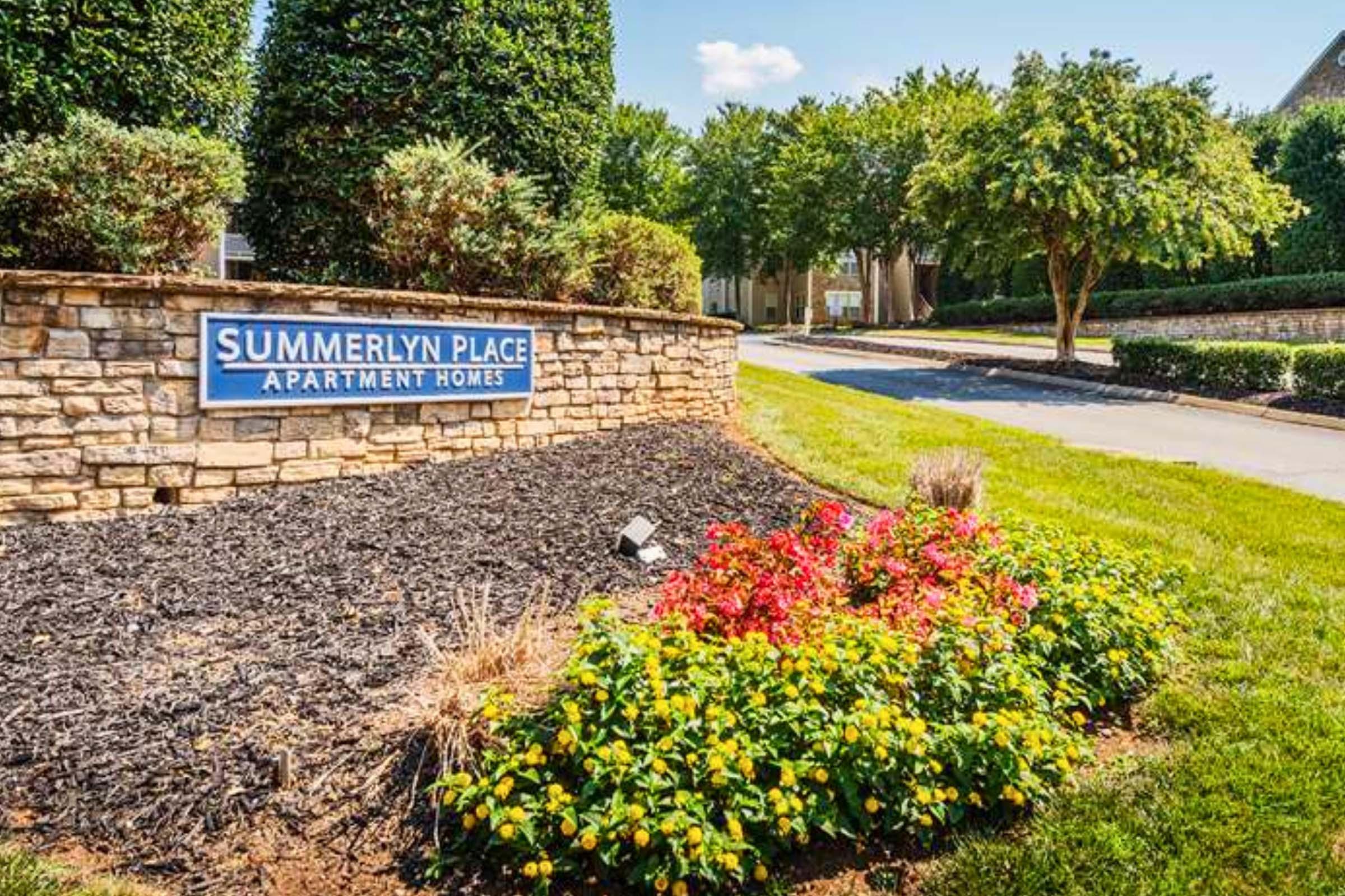 Sign for Summerlyn Place Apartment Homes, surrounded by landscaped greenery and colorful flower beds. The scene features a tidy pathway and trees under a bright blue sky, conveying a welcoming atmosphere.