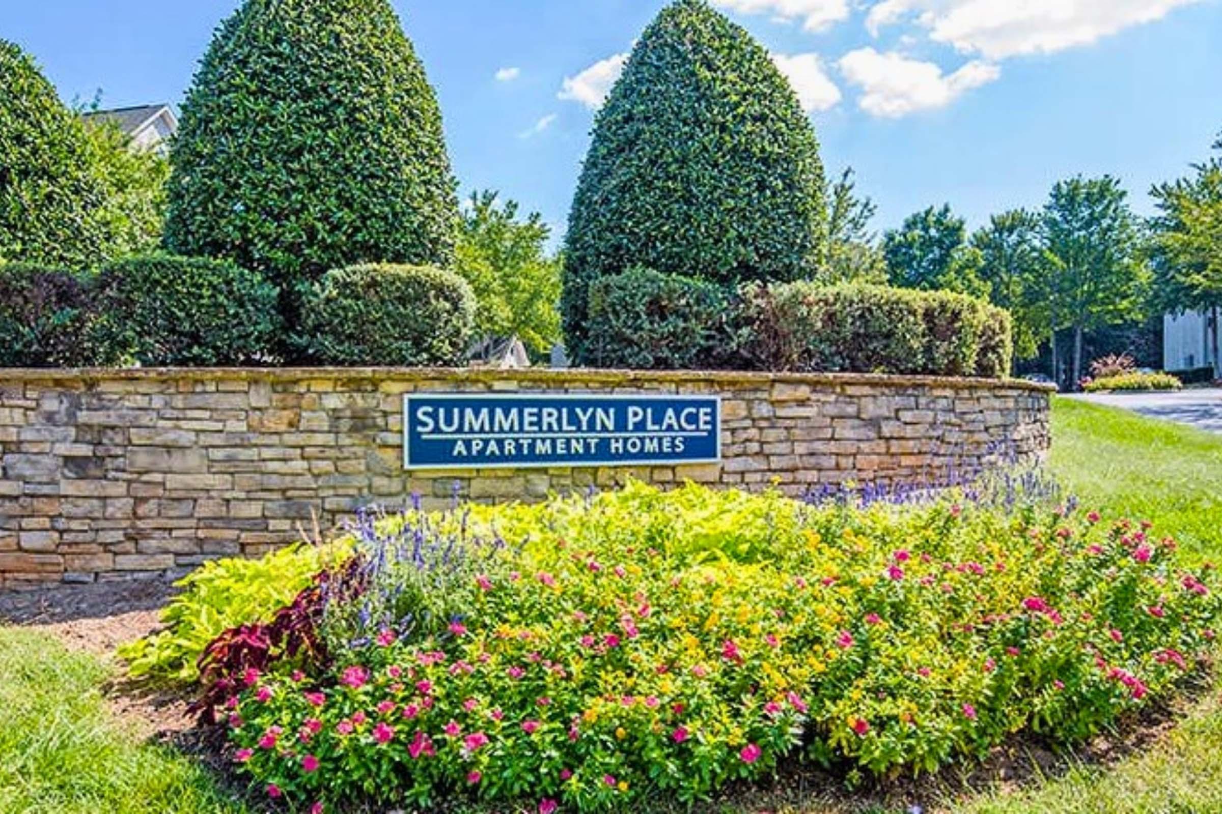 Sign for Summerlyn Place Apartment Homes surrounded by manicured hedges and colorful flower beds. Bright blue sky with a few clouds in the background, creating a welcoming atmosphere for residents and visitors.
