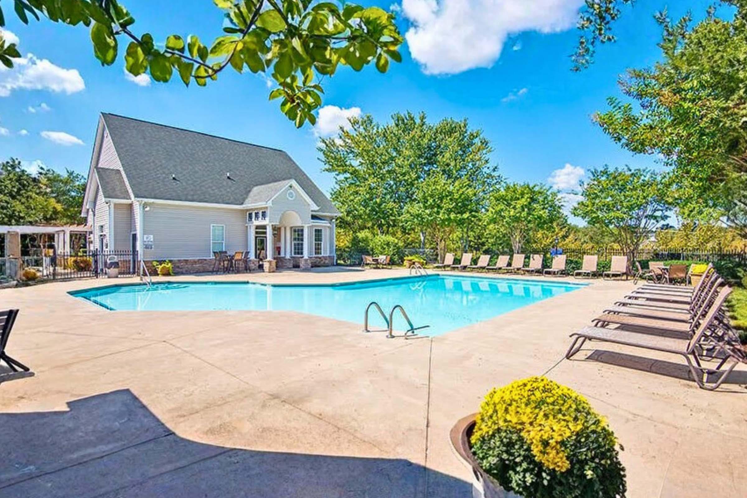 A sunny outdoor pool area featuring a clear blue pool surrounded by lounge chairs, with a well-maintained house and lush greenery in the background under a bright blue sky with fluffy clouds.