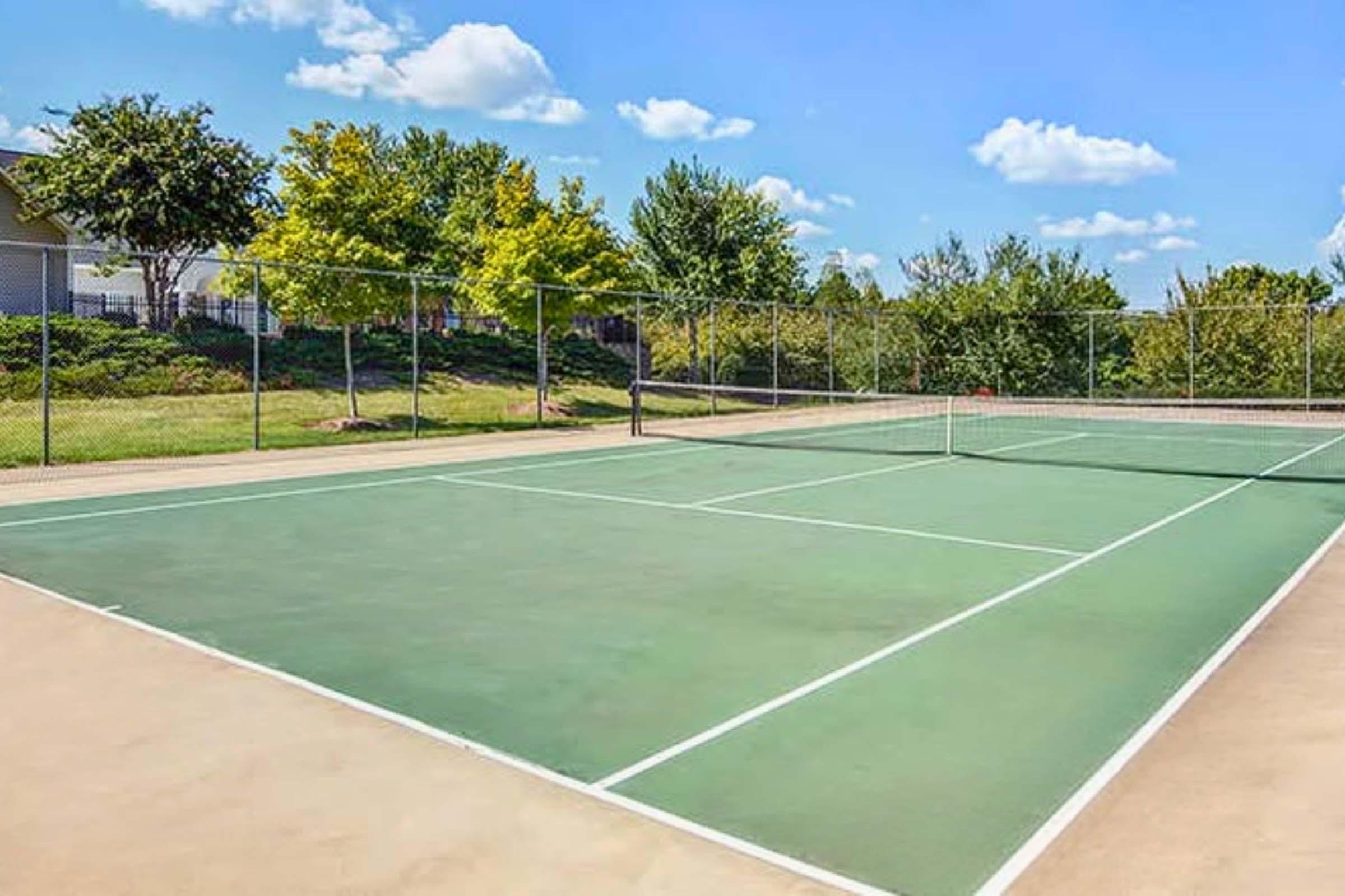 A well-maintained outdoor tennis court with a green surface, surrounded by a chain-link fence. Lush trees and greenery are visible in the background under a clear blue sky with fluffy white clouds. The court is empty, offering a serene setting for tennis.