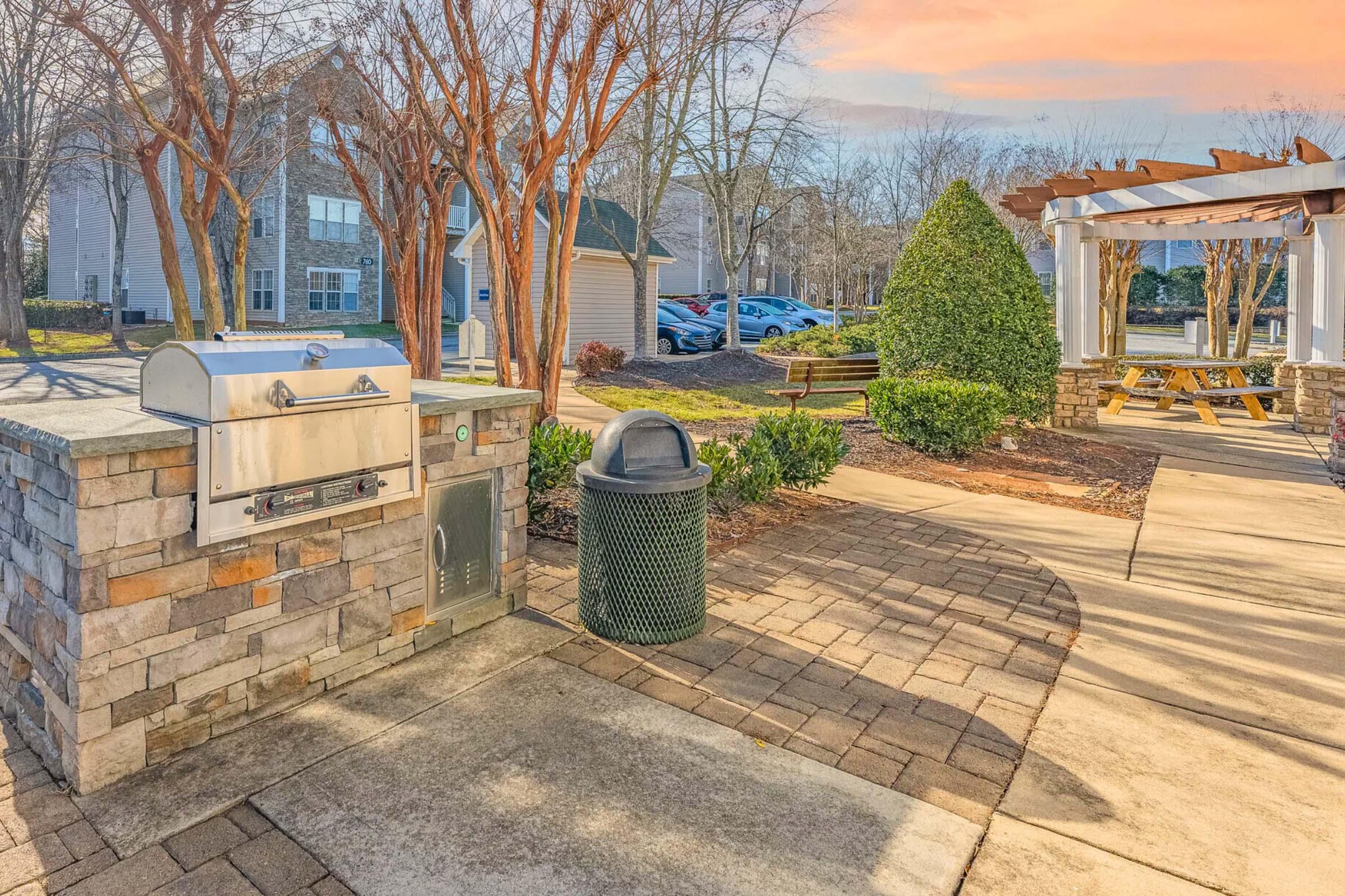 A communal outdoor area featuring a stone-built barbecue grill next to a green trash can. Surrounding the space are neatly landscaped bushes and trees, with a sunny sky and residential buildings visible in the background. A seating area under a pergola can be seen further down the path.