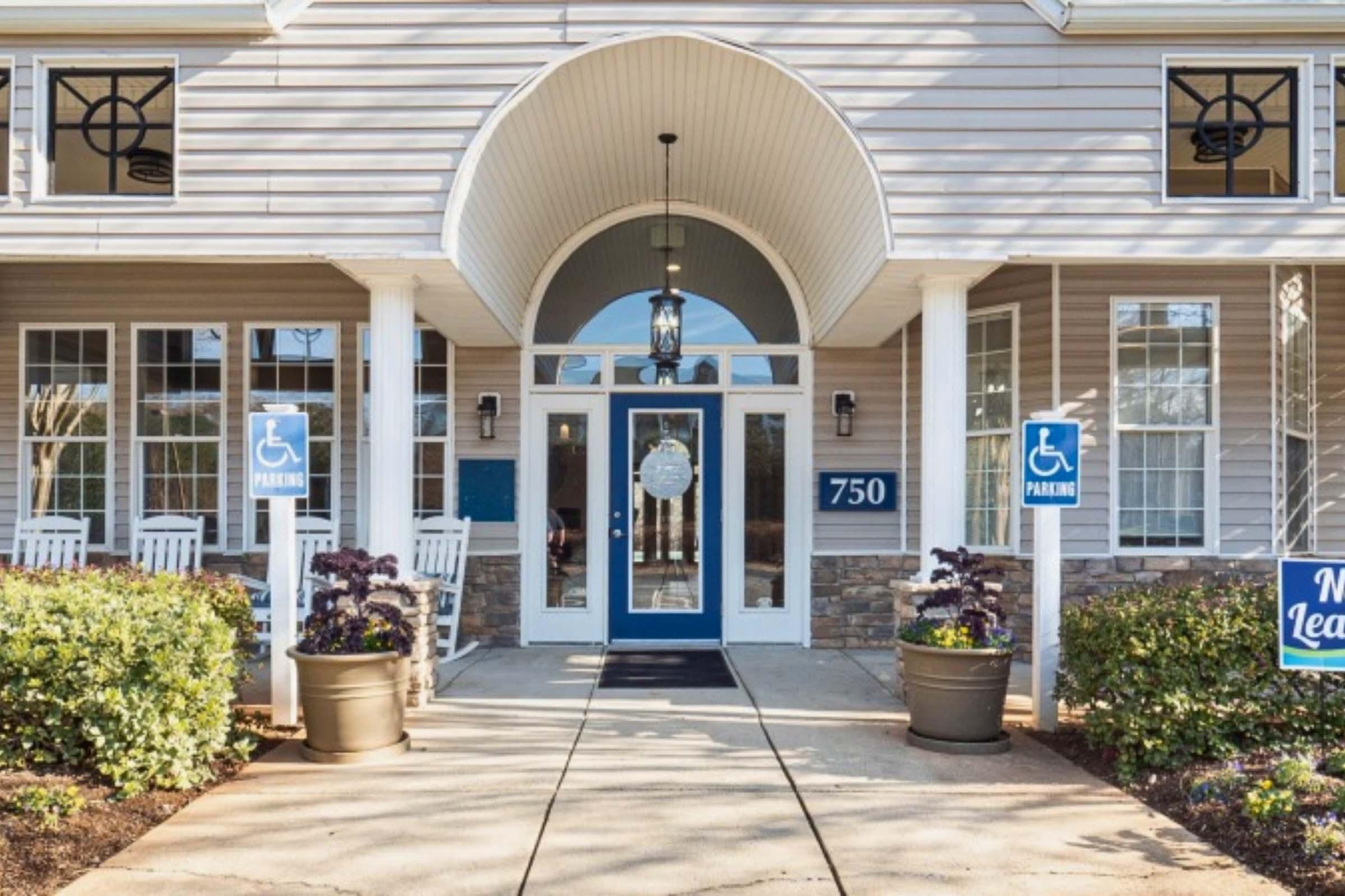 Entrance to a building featuring a large arched doorway and a blue door with a circular sign. There are two planters with purple flowers on either side. Signs indicate accessible parking and a "Now Leasing" advertisement. The façade is light-colored with stone accents and rocking chairs on the porch.