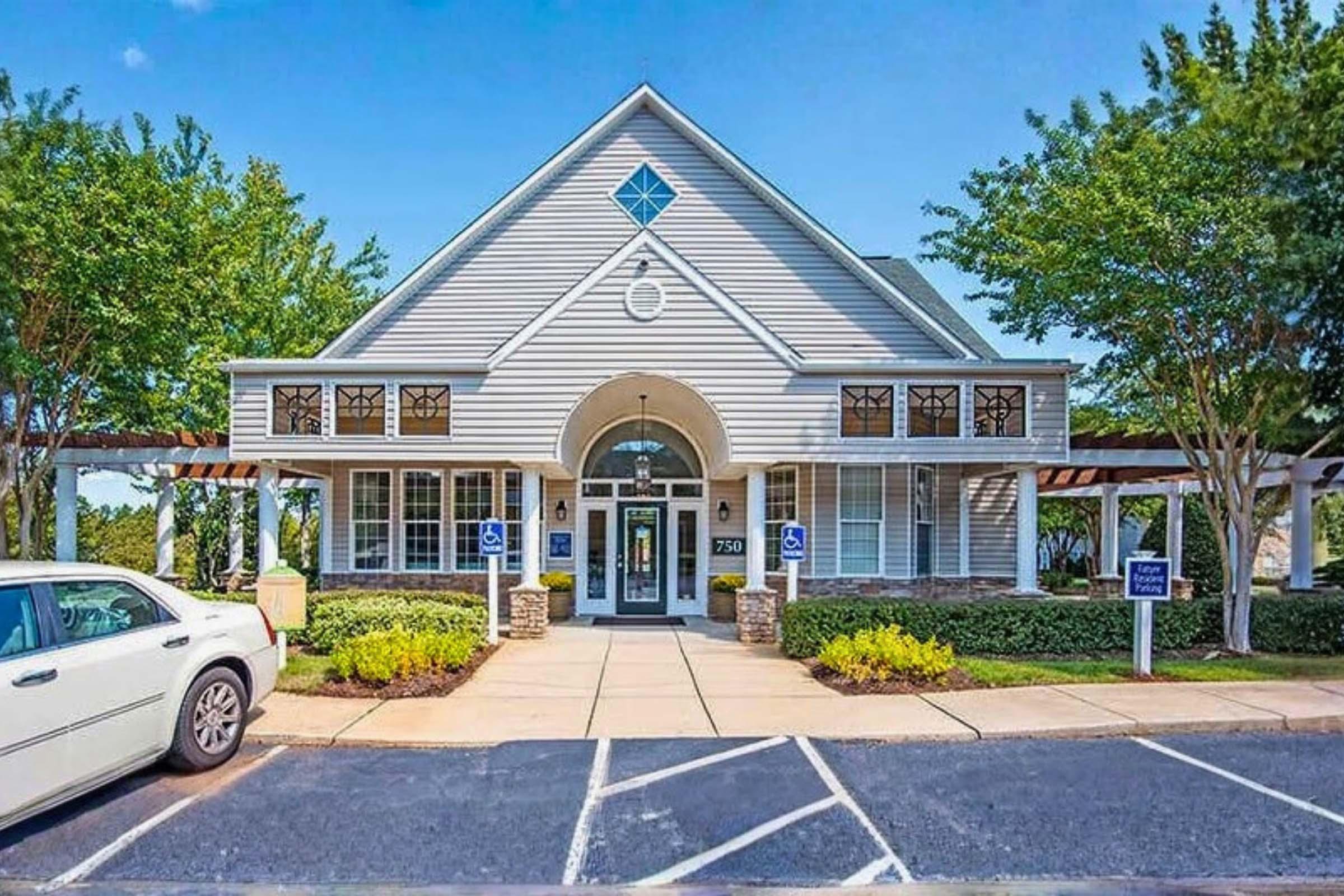 A modern, two-story building with a light gray exterior and a triangular rooftop. It features large windows with decorative panes, a central entrance with a round arch, and landscaping that includes trees and shrubs. There is a parking area in front with a white car parked.