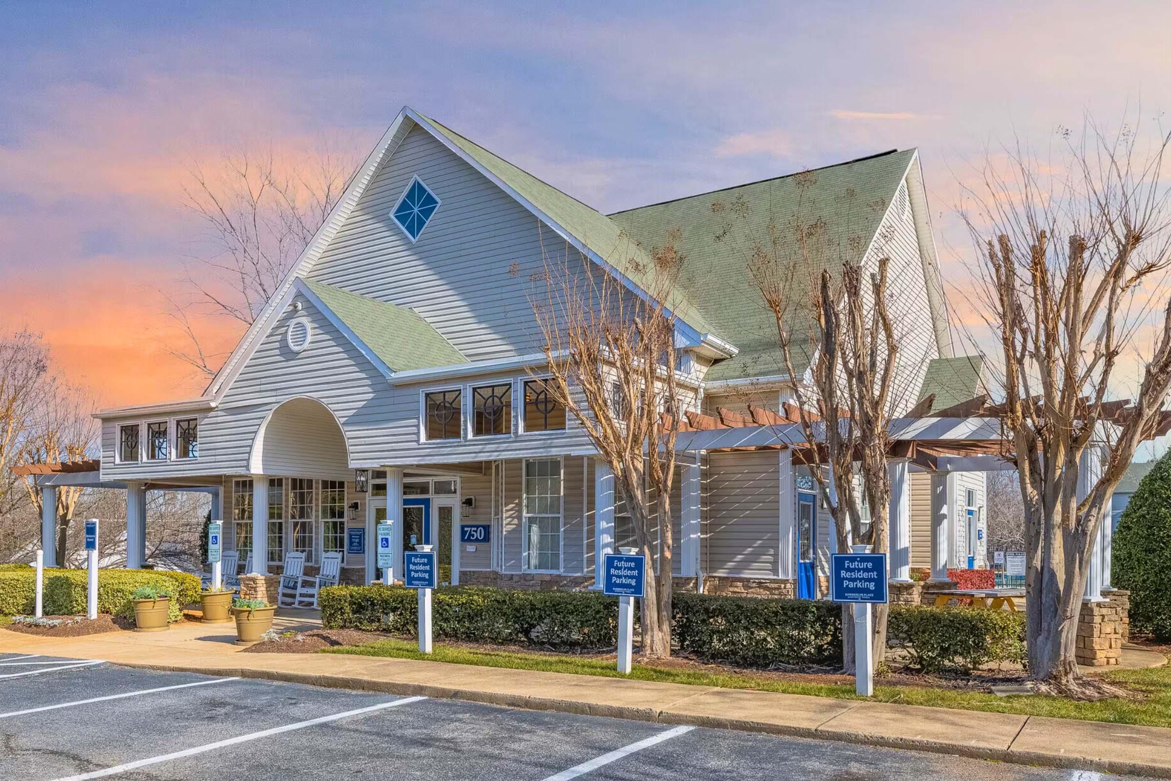 A charming light gray building with a green roof, featuring large windows and a decorative archway entrance. The exterior is complemented by seasonal trees and signs indicating reserved parking areas. The sky in the background displays soft pastel colors at sunset.
