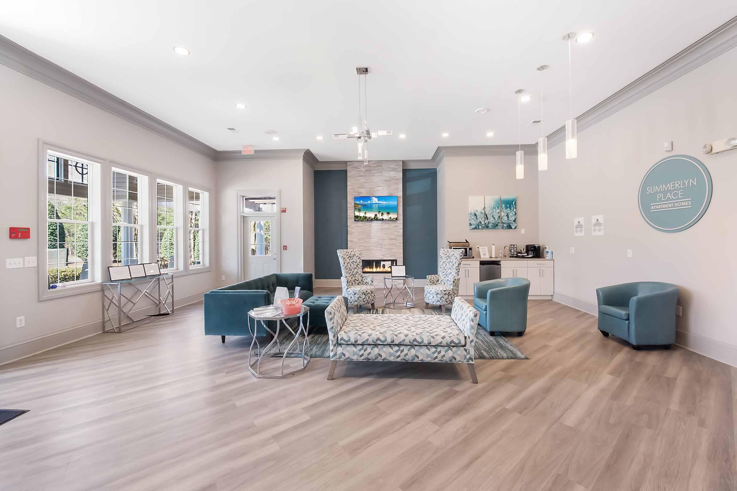 Modern lobby of an apartment complex featuring comfortable seating arrangements, including teal chairs and a patterned couch. The space is well-lit with large windows, a contemporary coffee station, and decorative artwork on the walls. A TV mounted above the fireplace enhances the welcoming atmosphere.