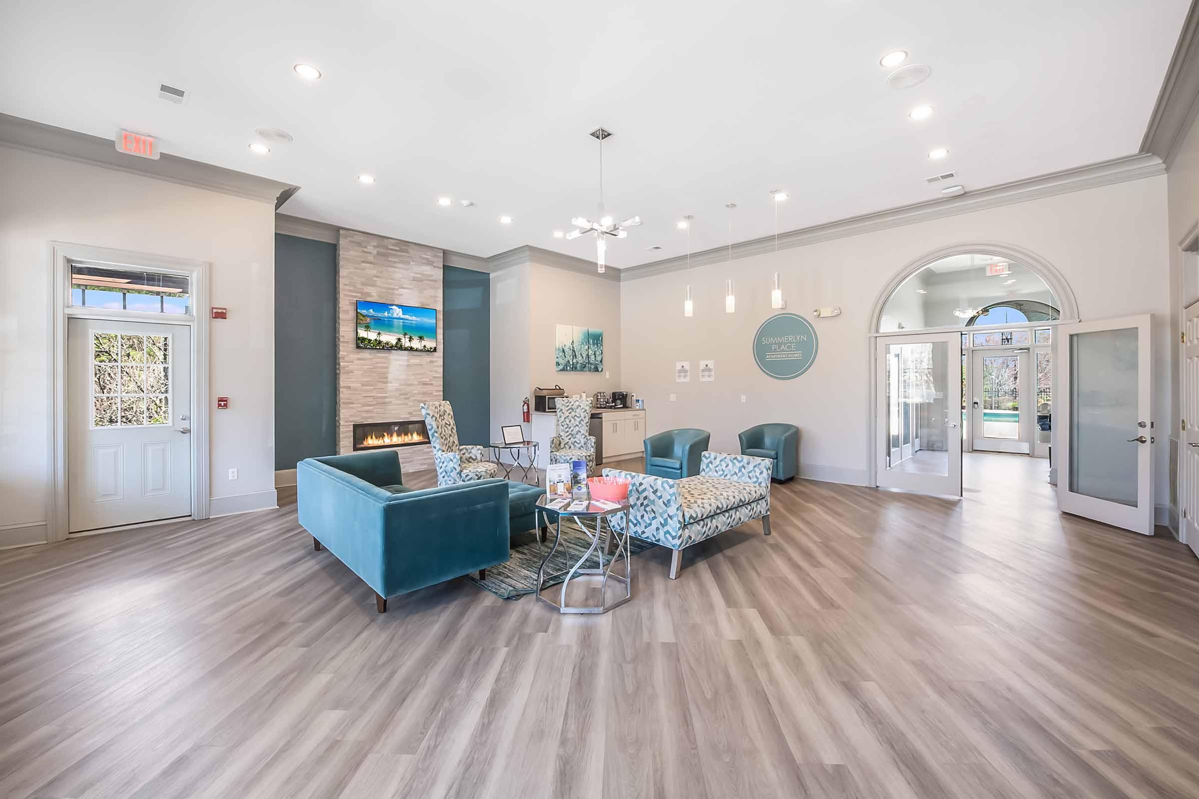 Interior view of a modern lobby featuring stylish furniture, including blue armchairs and a patterned sofa. A stone fireplace adds warmth, and large windows allow natural light. The room is decorated with artwork and has a welcoming atmosphere. Bright overhead lights and light wood flooring enhance the space.