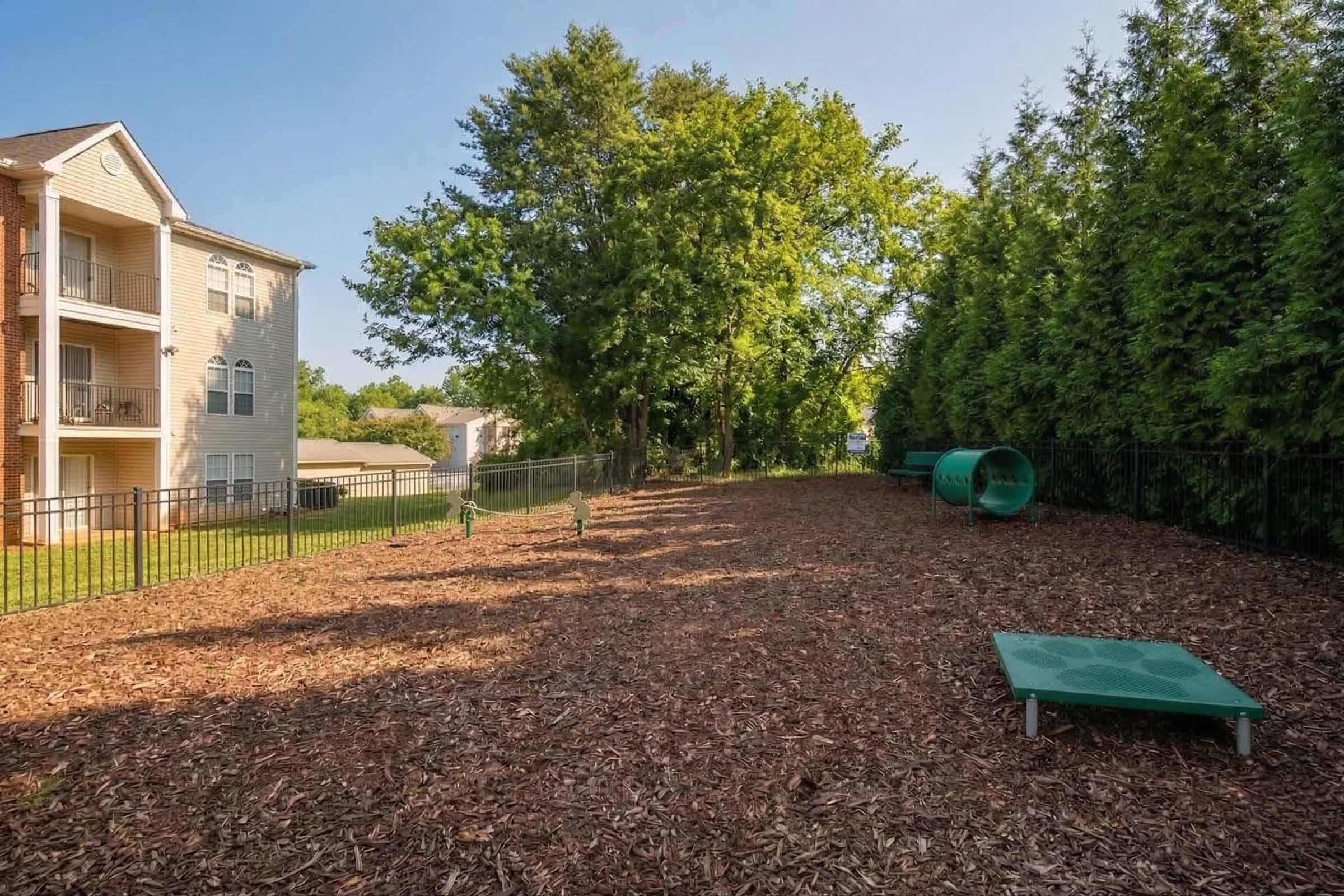 A spacious outdoor area with a wood chip ground, surrounded by a black fence and tall greenery. In the background, there are two residential buildings and a play area featuring two green tube slides and raised platforms for pets. The scene is bright and sunny, showcasing an inviting recreational space.