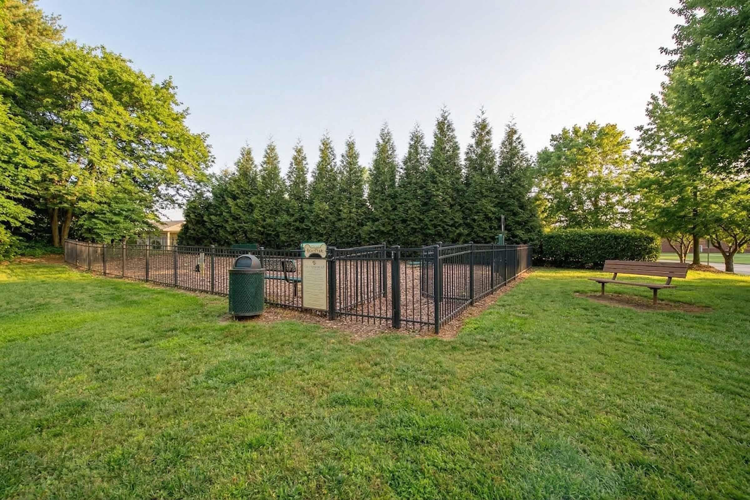 A fenced dog park featuring a grassy area surrounded by trees, with a bench for seating and a trash bin nearby. The park has a gravel surface and a sign at the entrance. Ideal space for dogs to play and socialize in a safe environment.