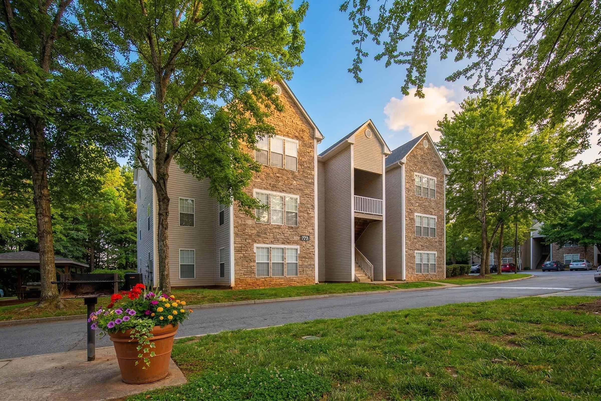 A three-story residential building with a mix of stone and siding exterior, surrounded by green trees. A flower pot with colorful blooms is in the foreground, and a parking area is visible alongside the building. The sky is clear with a few clouds.