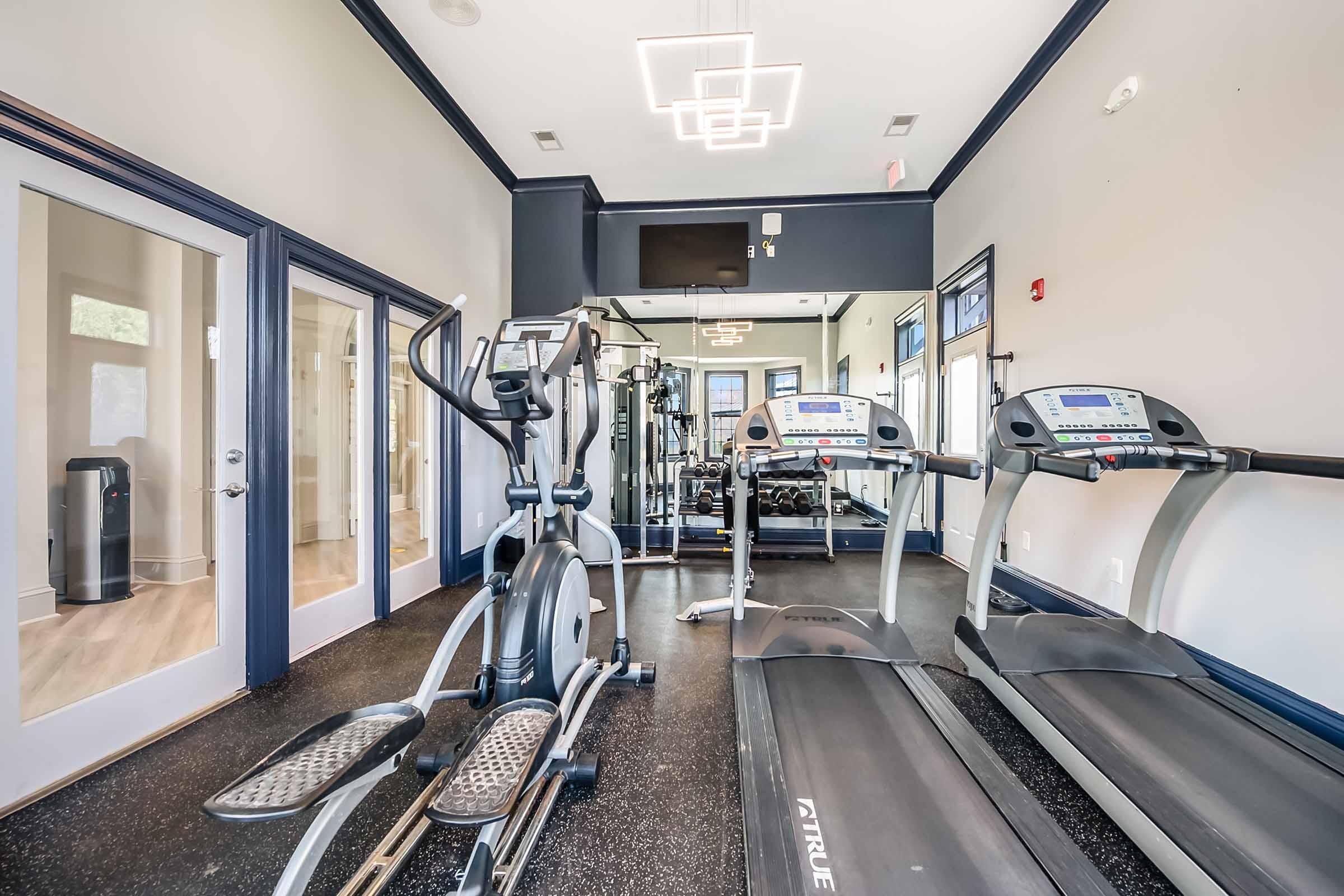 Interior view of a fitness room featuring two treadmills, an elliptical machine, and a weight machine. Large windows allow natural light to fill the space. The floor is dark and there are modern light fixtures on the ceiling, creating a bright and inviting atmosphere for exercise.