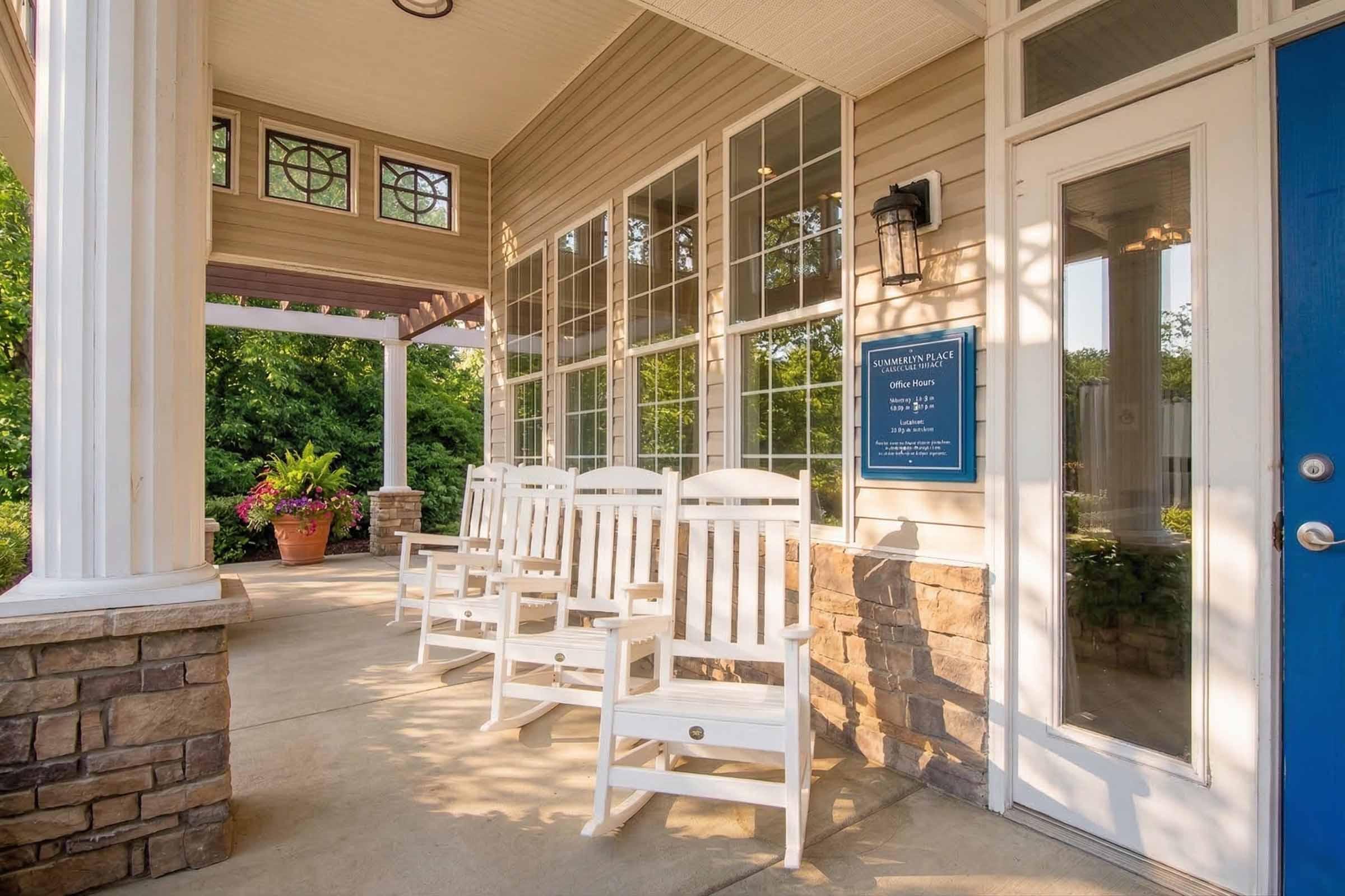 A welcoming entrance featuring a porch with several white rocking chairs arranged neatly. The porch has large windows allowing natural light and is adorned with potted flowers. A blue door leads into the building, accompanied by a sign mounted on the wall.