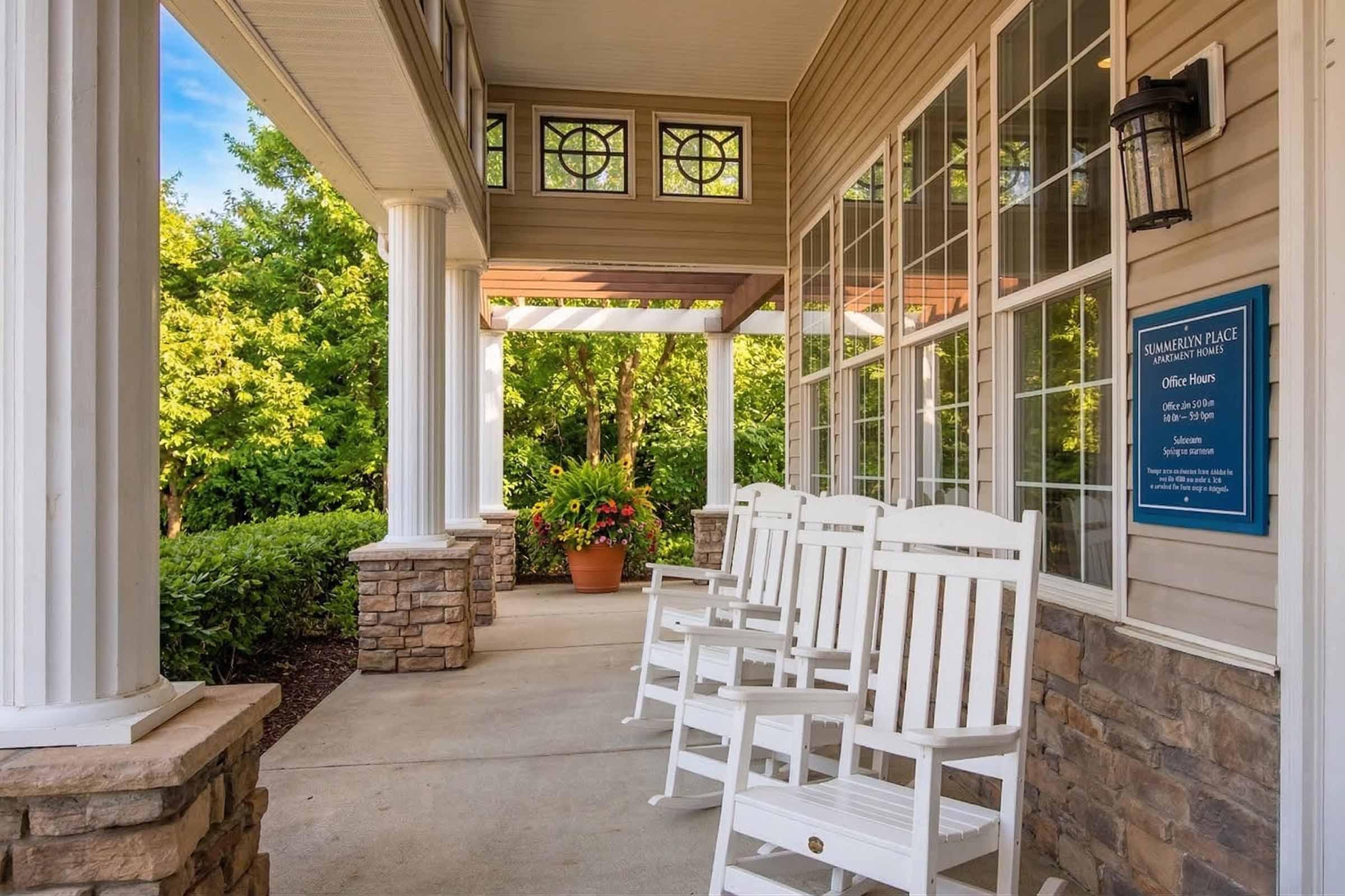 A welcoming porch with white rocking chairs and stone pillars, surrounded by lush greenery. The area is bright and inviting, featuring large windows and a sign indicating the office hours for a place called "Summerton Place."