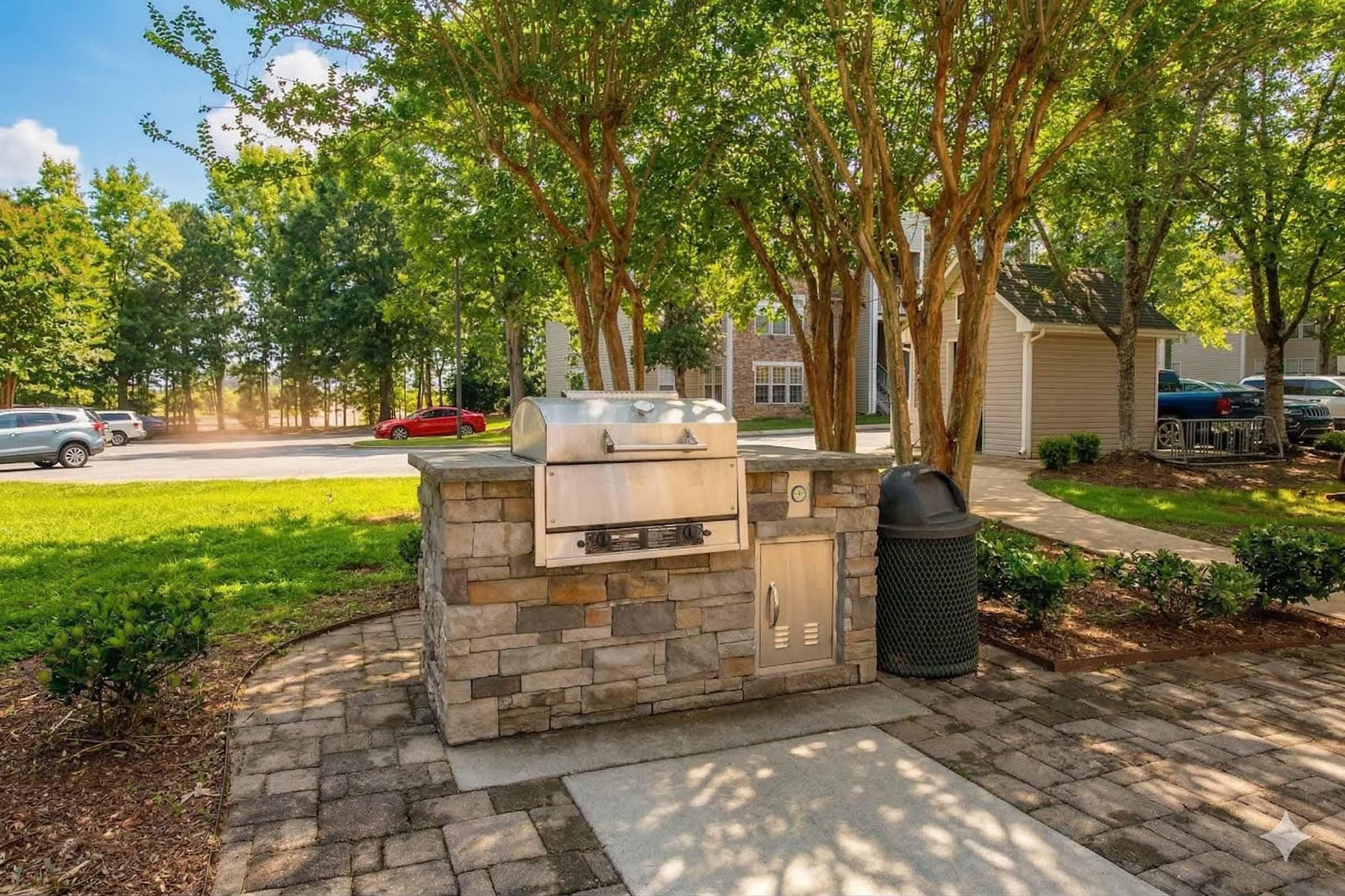 Outdoor grilling station featuring a stainless steel grill mounted on a stone counter, surrounded by well-maintained greenery and trees. A trash can is positioned nearby, and a paved pathway leads to the grill area. Bright blue sky and distant cars create a welcoming ambiance in a residential setting.