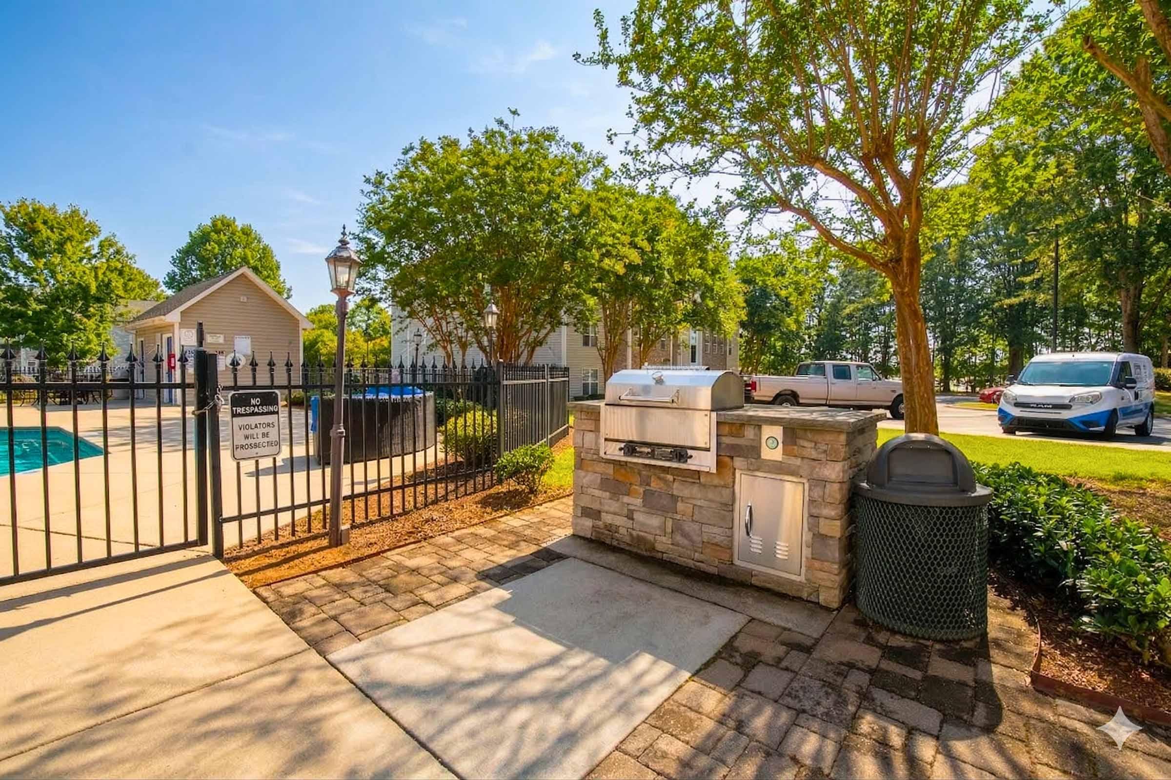 Grill area by a swimming pool, surrounded by a black gate and greenery. A stone grill is located next to a trash can, with a clear blue sky above. Nearby, residential buildings and parked vehicles are visible, creating a welcoming outdoor space.