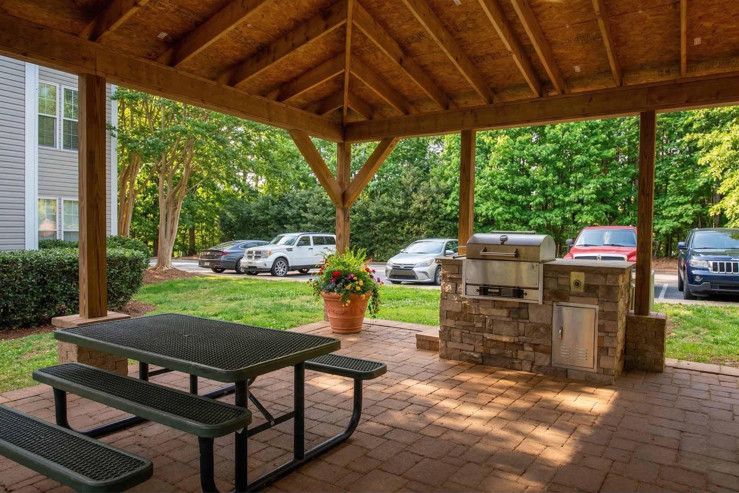 A shaded outdoor space featuring a wooden pavilion with a picnic table and a stone grill station. Lush greenery surrounds the area, with parked cars in the background. The setting is ideal for gatherings or barbecues in a serene environment.