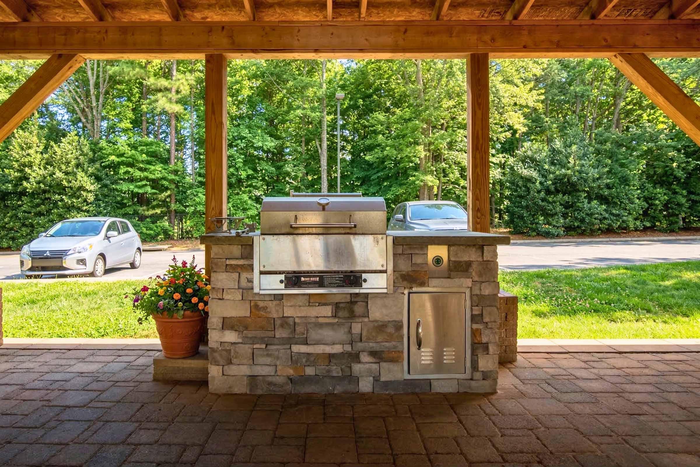 Outdoor grilling area featuring a stone grill setup under a wooden pavilion, with a silver grill, a storage compartment, and a flower pot with vibrant flowers. In the background, there are trees and two parked cars visible.