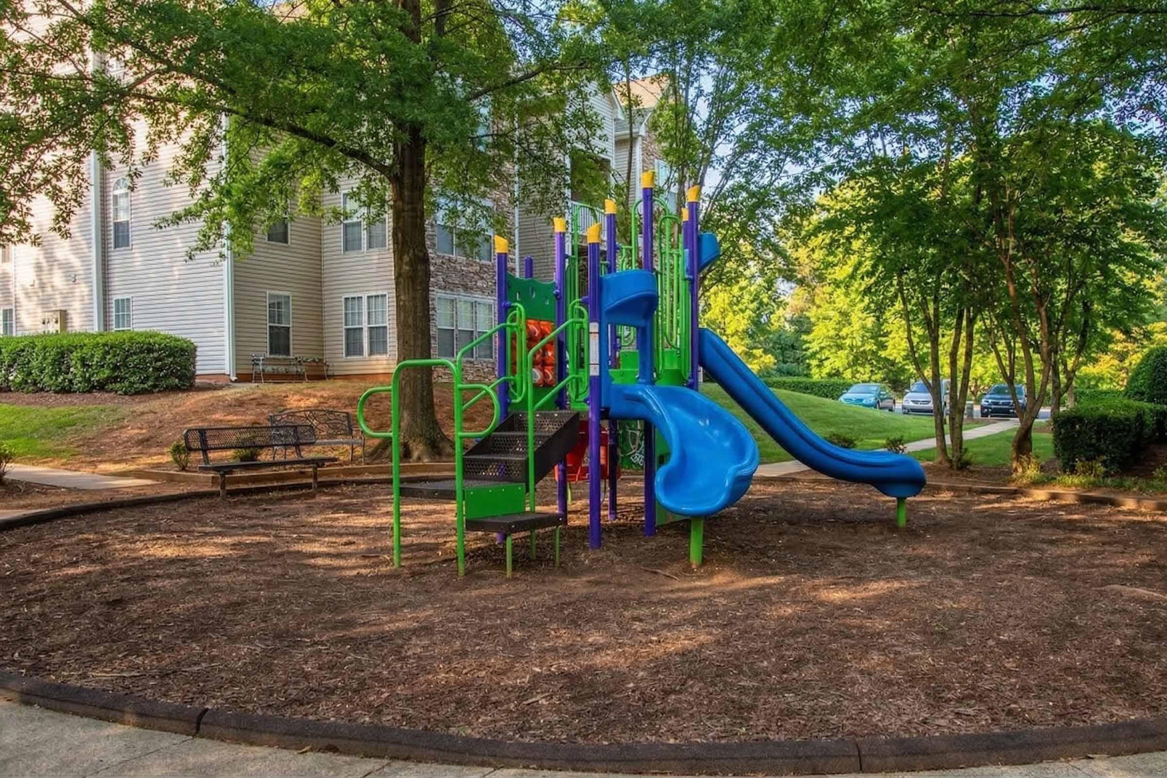 Colorful playground equipment featuring slides and climbing structures, surrounded by a grassy area with trees and benches. In the background, there are residential buildings and parked cars. The playground is designed for children's recreation in a suburban setting.