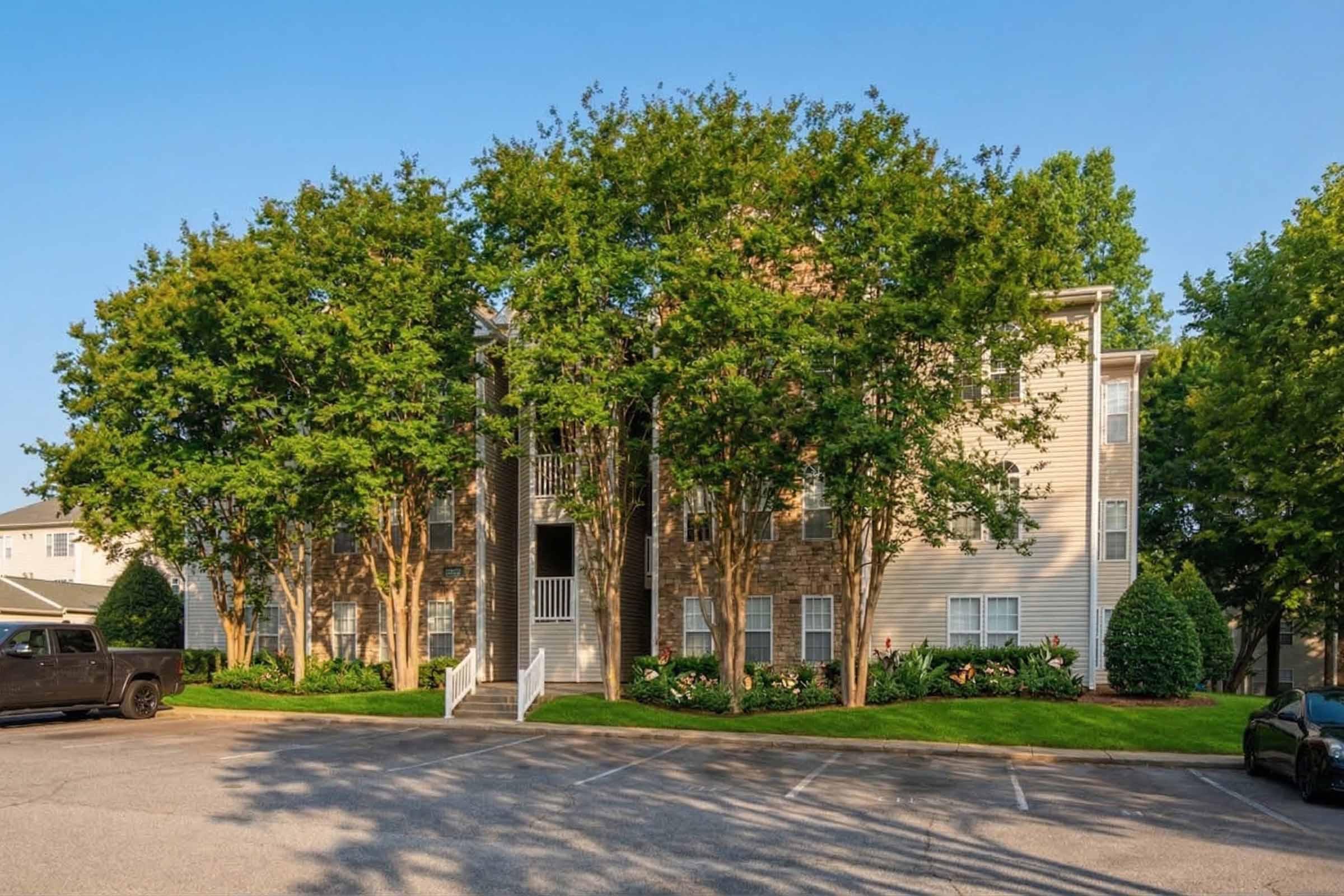 A three-story residential apartment building with a mixture of stone and siding exterior, surrounded by well-maintained trees and shrubs. The building features a central entrance with stairs and balconies. A parking area in the foreground includes several spaces and a black pickup truck. Clear blue sky overhead.