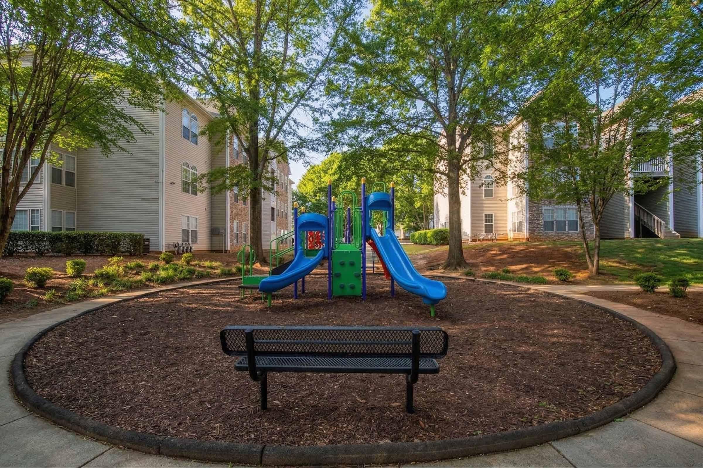 A playground featuring colorful slides and climbing structures is situated in a landscaped area surrounded by trees. A black bench is positioned nearby on a paved pathway, providing a spot for relaxation. Residential buildings are visible in the background.