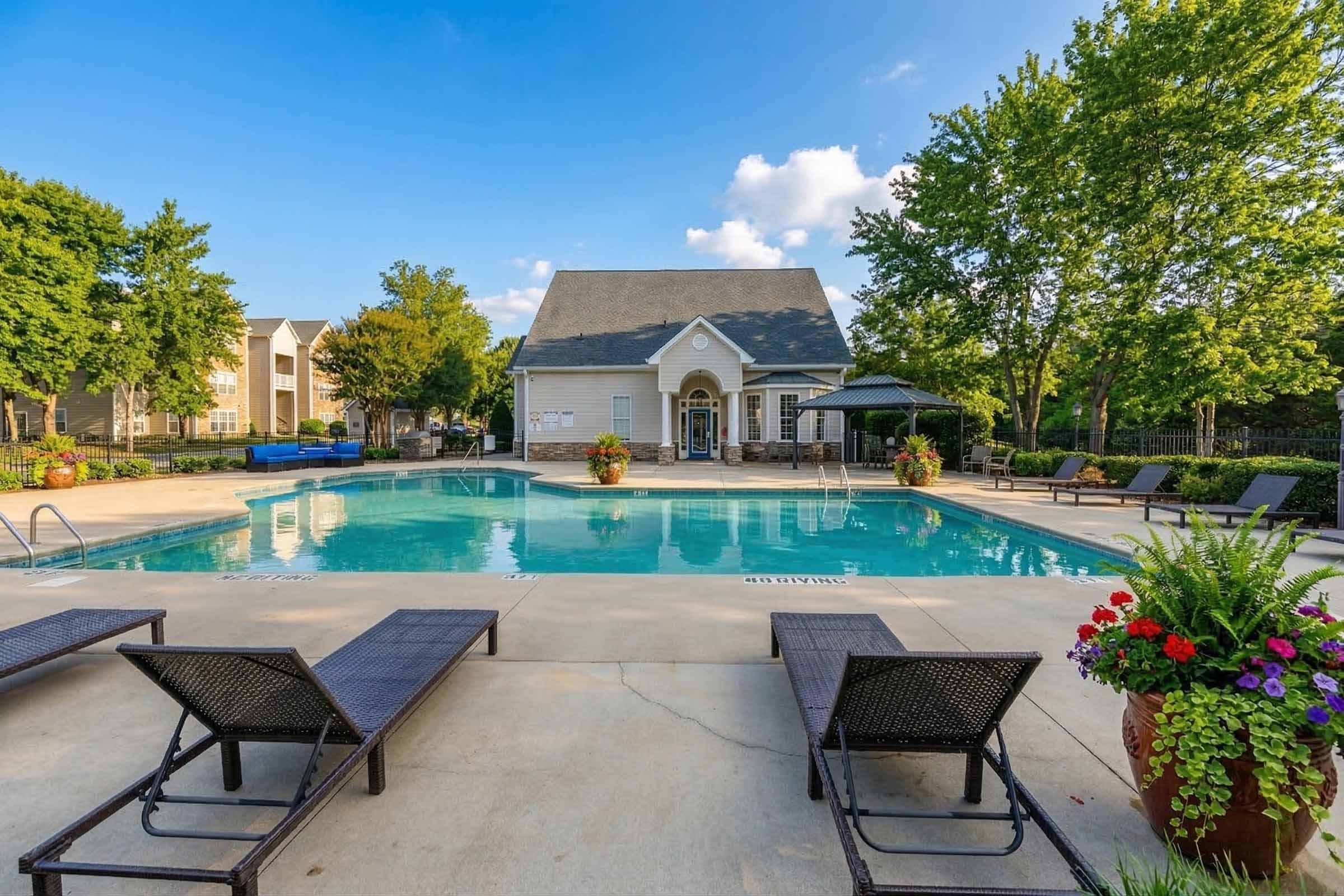 A clear blue swimming pool surrounded by lounge chairs, vibrant flowers, and trees. In the background, there is a clubhouse with a gabled roof. The sky is partly cloudy, creating a relaxed outdoor atmosphere ideal for leisure.