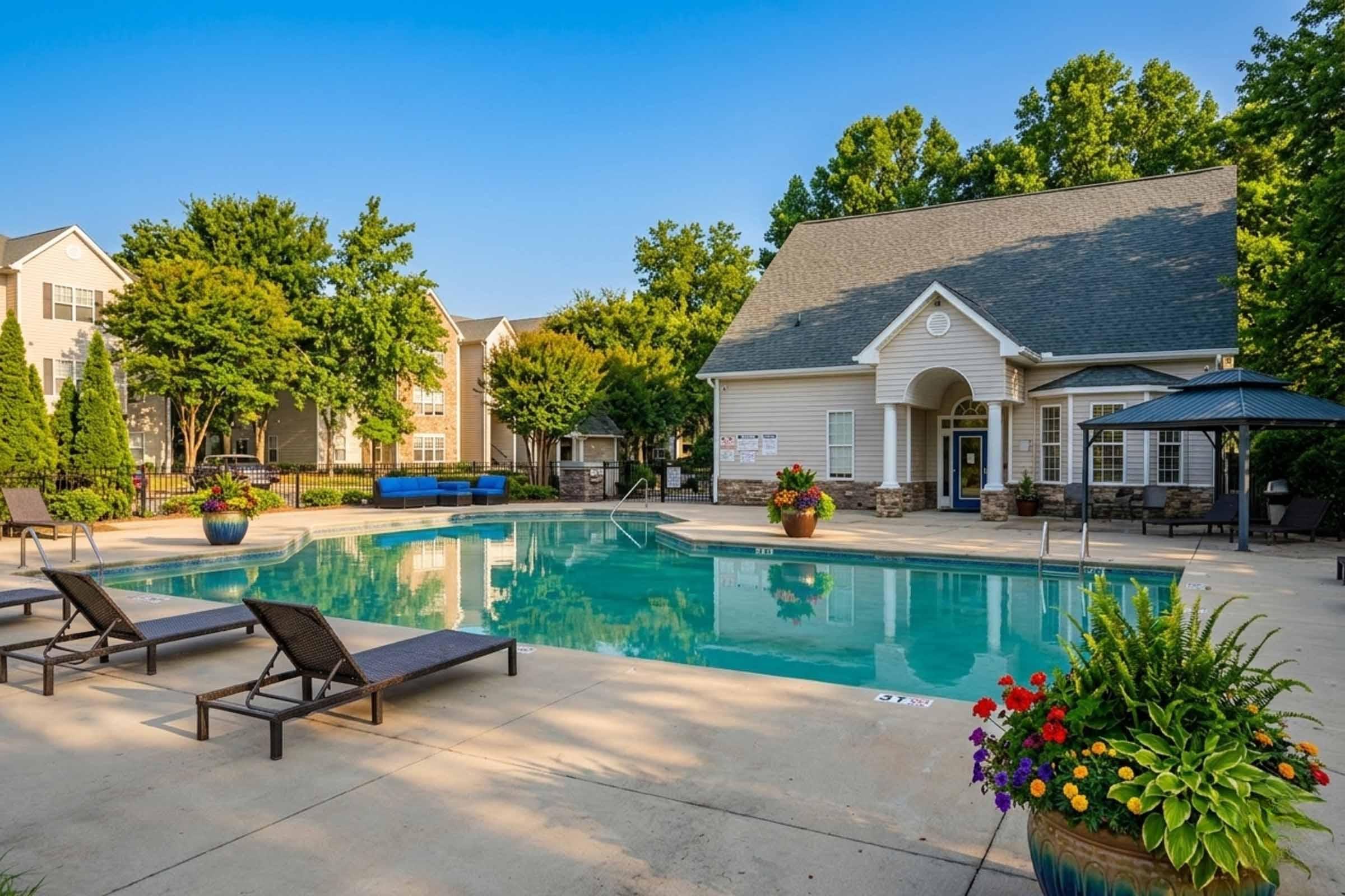 A serene outdoor swimming pool area surrounded by lounge chairs and colorful flower pots. There are trees in the background and a clubhouse entrance with a welcoming design. The sky is clear and blue, creating a bright and inviting atmosphere.