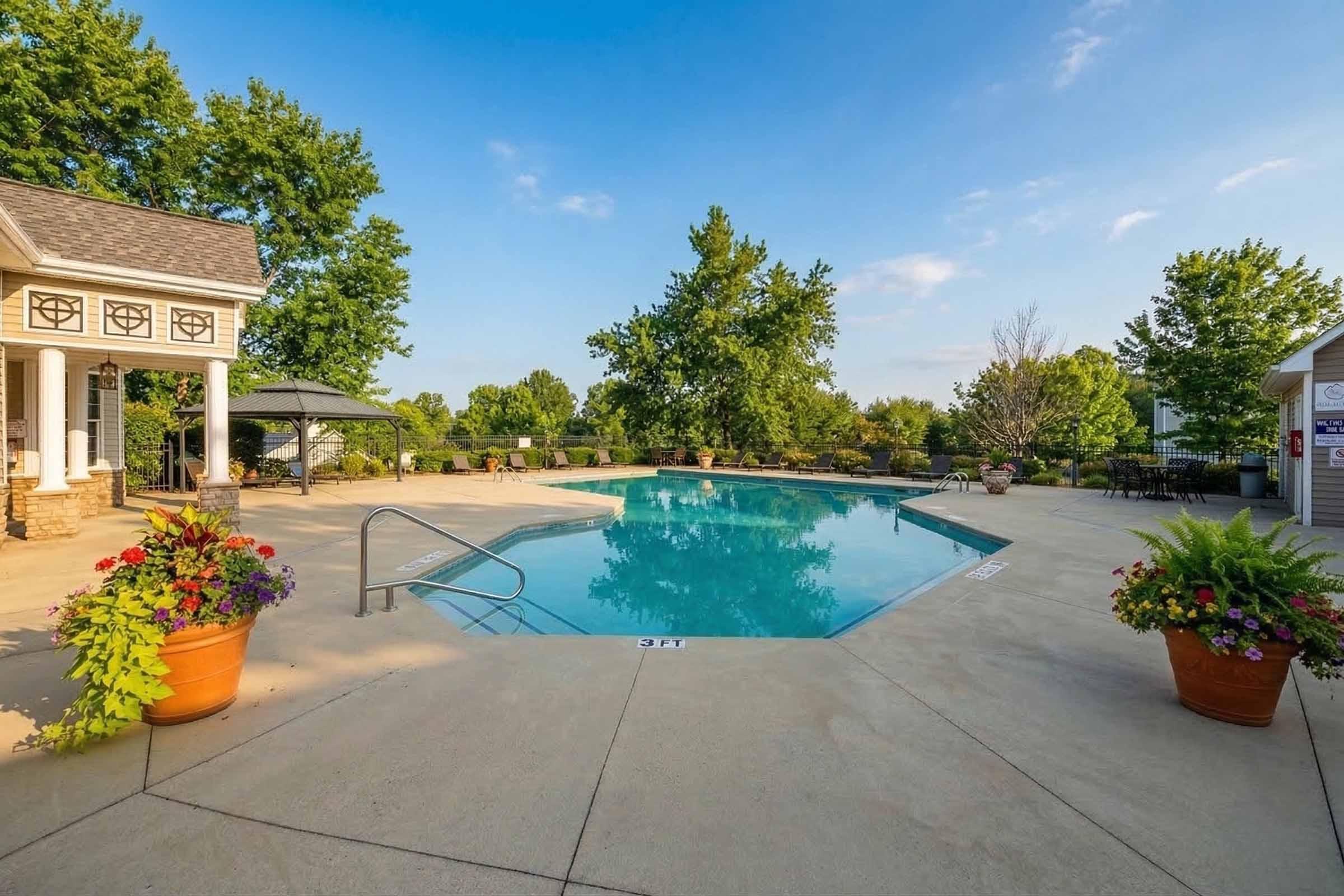 A serene outdoor swimming pool surrounded by lush greenery. The pool features a smooth, clear surface and is framed by well-maintained concrete. Potted plants with vibrant flowers are placed around the pool area, enhancing the inviting atmosphere. A gazebo and outdoor seating area are visible in the background under a bright blue sky.