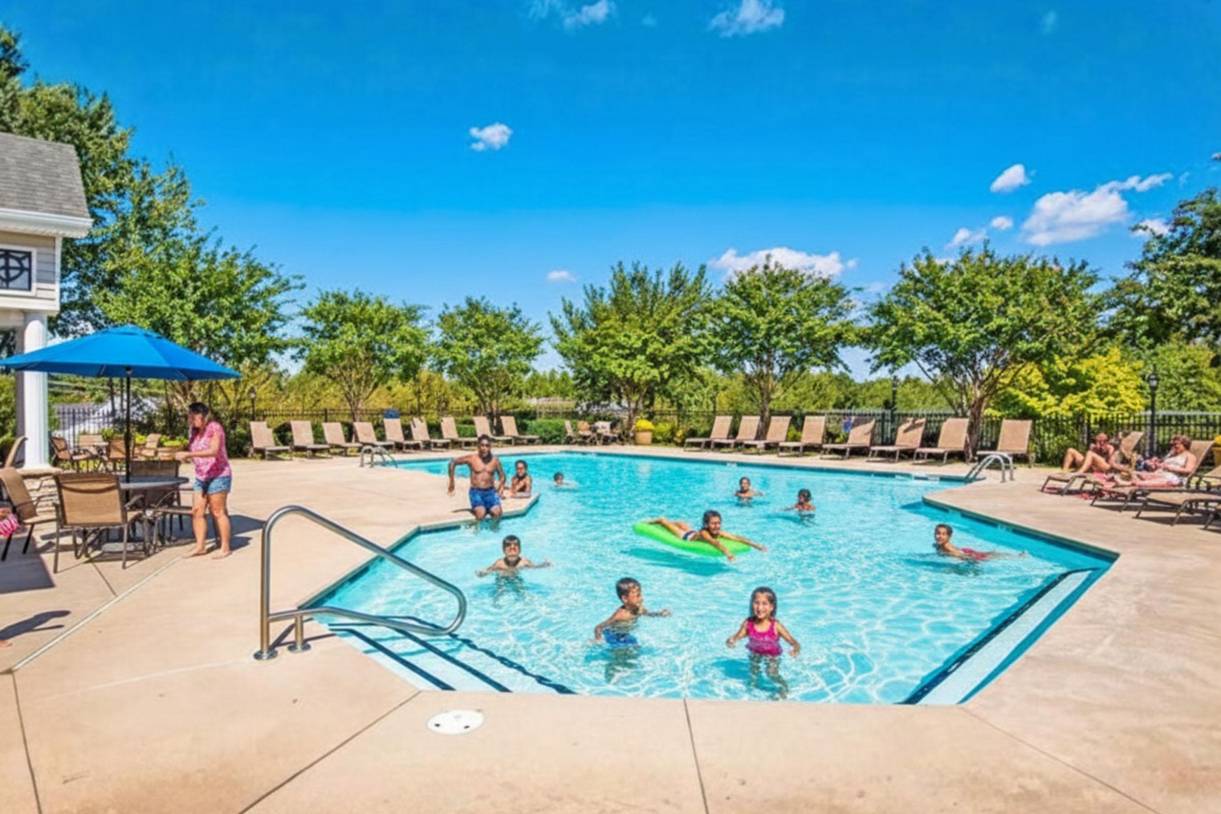 A vibrant outdoor swimming pool scene on a sunny day. Children and adults are enjoying the water, with some playing and others lounging on poolside chairs. Lush green trees surround the area, creating a relaxing atmosphere. Blue skies and fluffy clouds add to the cheerful setting.