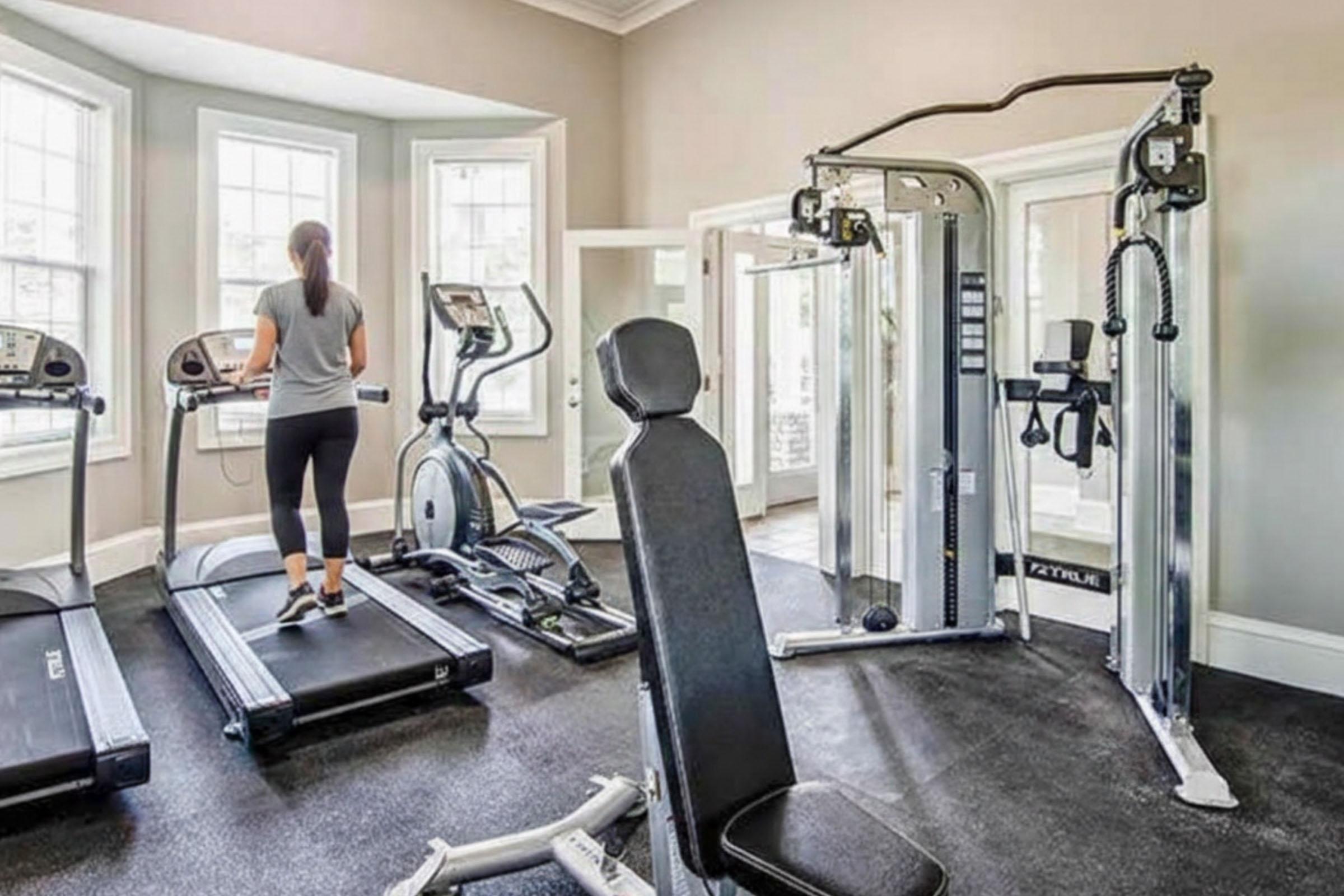 A modern fitness room featuring a treadmill, an elliptical machine, and a multi-gym station. A person in workout attire is walking on the treadmill, with a black weight bench positioned nearby. The room is well-lit with windows, creating a bright and inviting atmosphere for exercise.