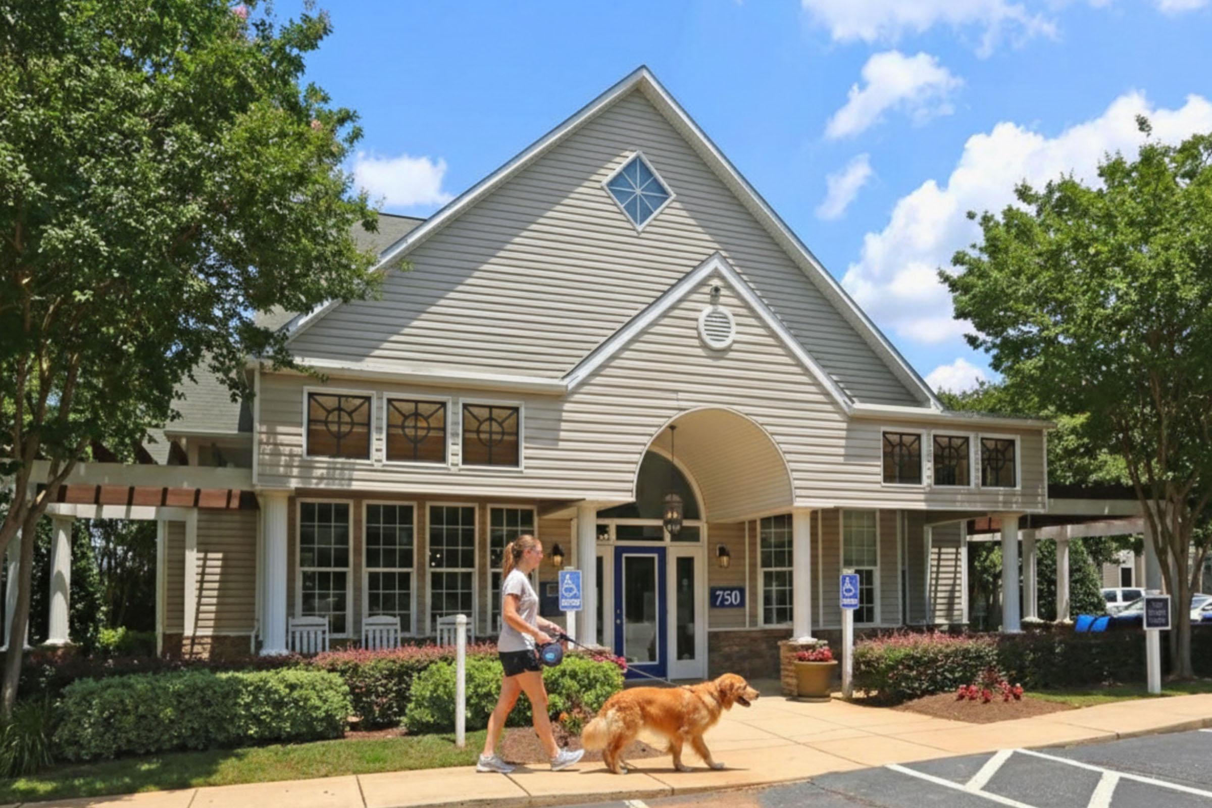 A person walking a golden retriever dog on a pathway in front of a light-colored building with large windows, surrounded by green trees and shrubs, under a blue sky with clouds. The building features accessible parking signs and a welcoming entrance.