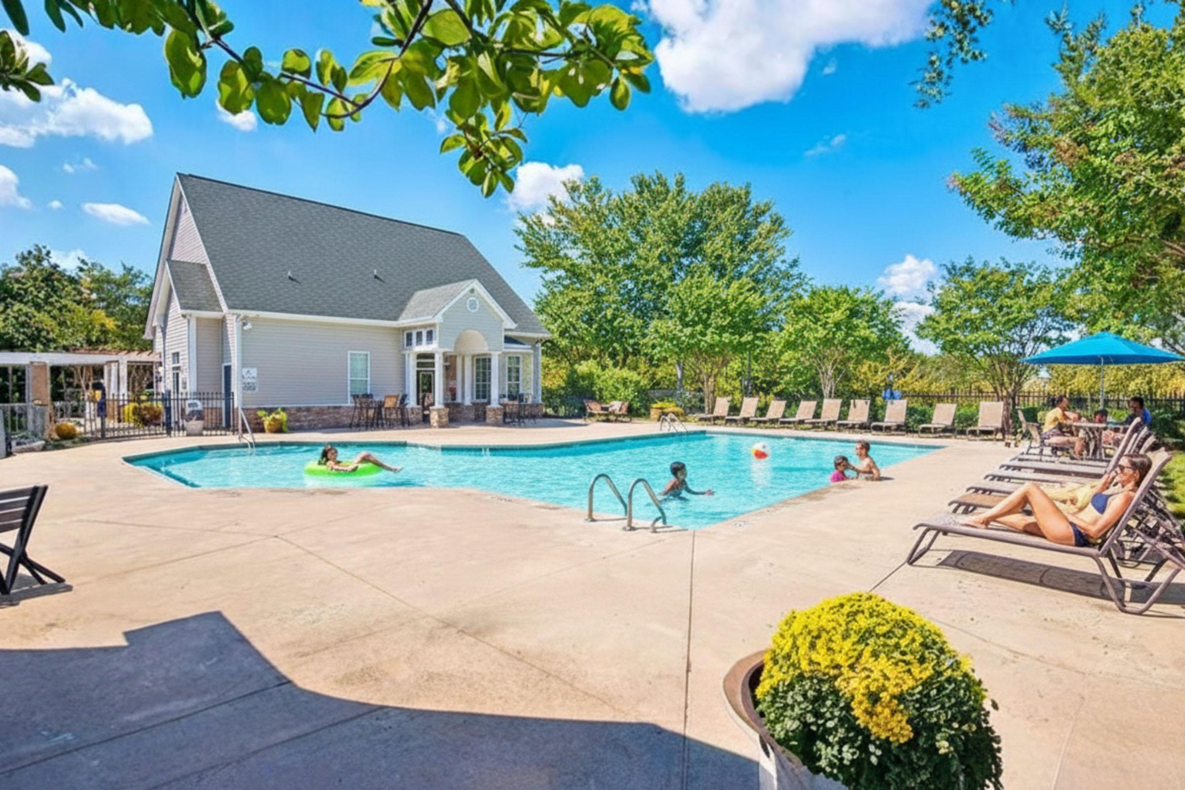 A sunny outdoor pool area featuring a clear blue swimming pool surrounded by lounging chairs. Several people are enjoying the water and sunbathing, while lush greenery and a charming house are in the background. Umbrellas provide shade, creating a relaxing atmosphere.