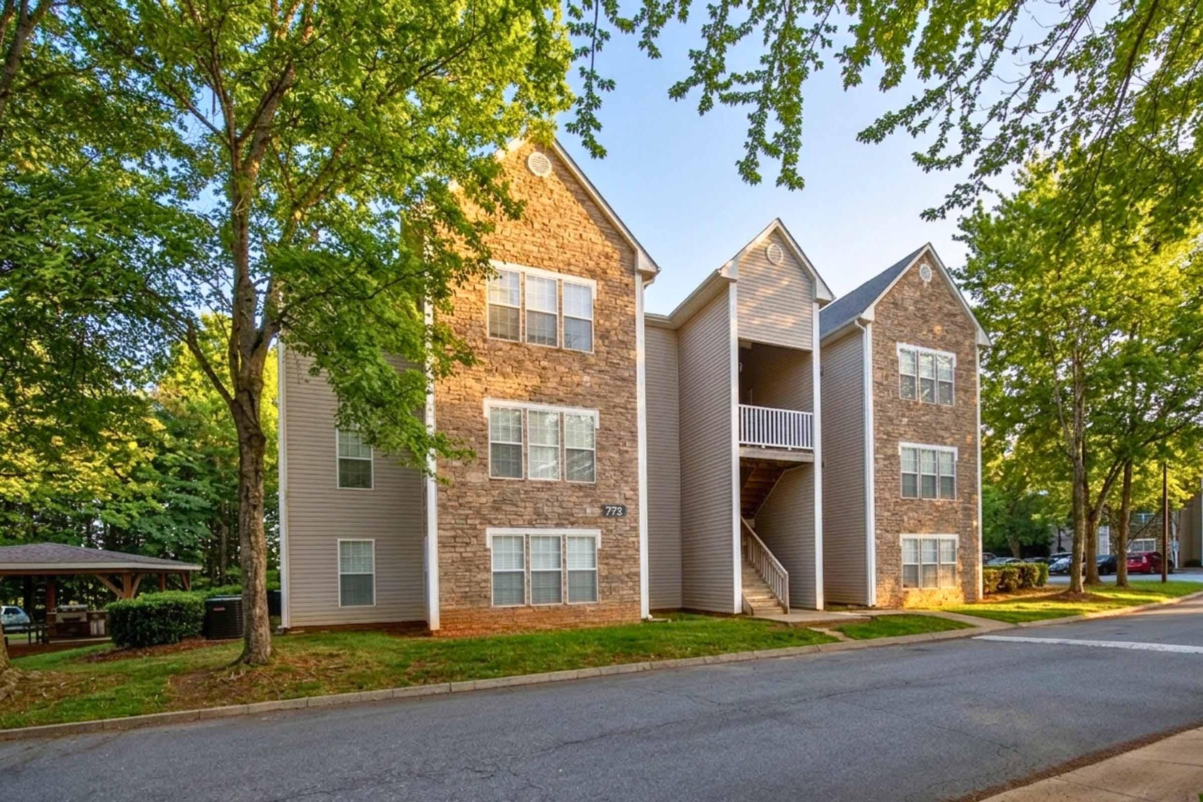 Three-story residential building with a combination of stone and siding exterior, featuring multiple windows and a staircase leading to the second level. Surrounding the building are green trees and a well-maintained lawn, with a street visible in front.