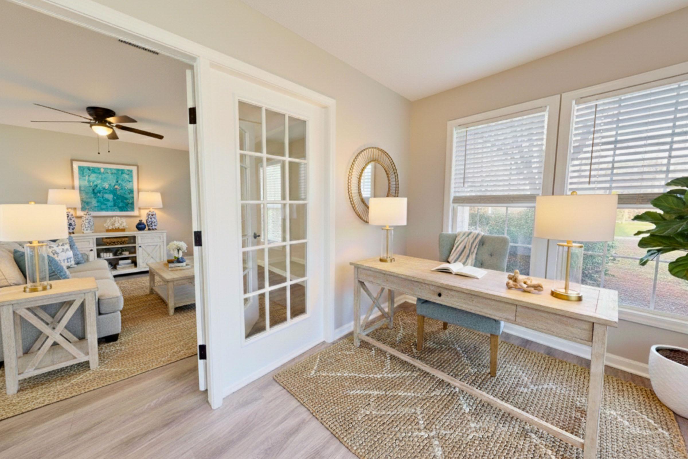 A bright, inviting home office featuring a wooden desk with a book and decorative items, surrounded by large windows with blinds. A cozy seating area with two blue armchairs is visible, along with a ceiling fan and a round mirror on the wall. A potted plant adds a touch of greenery to the space.