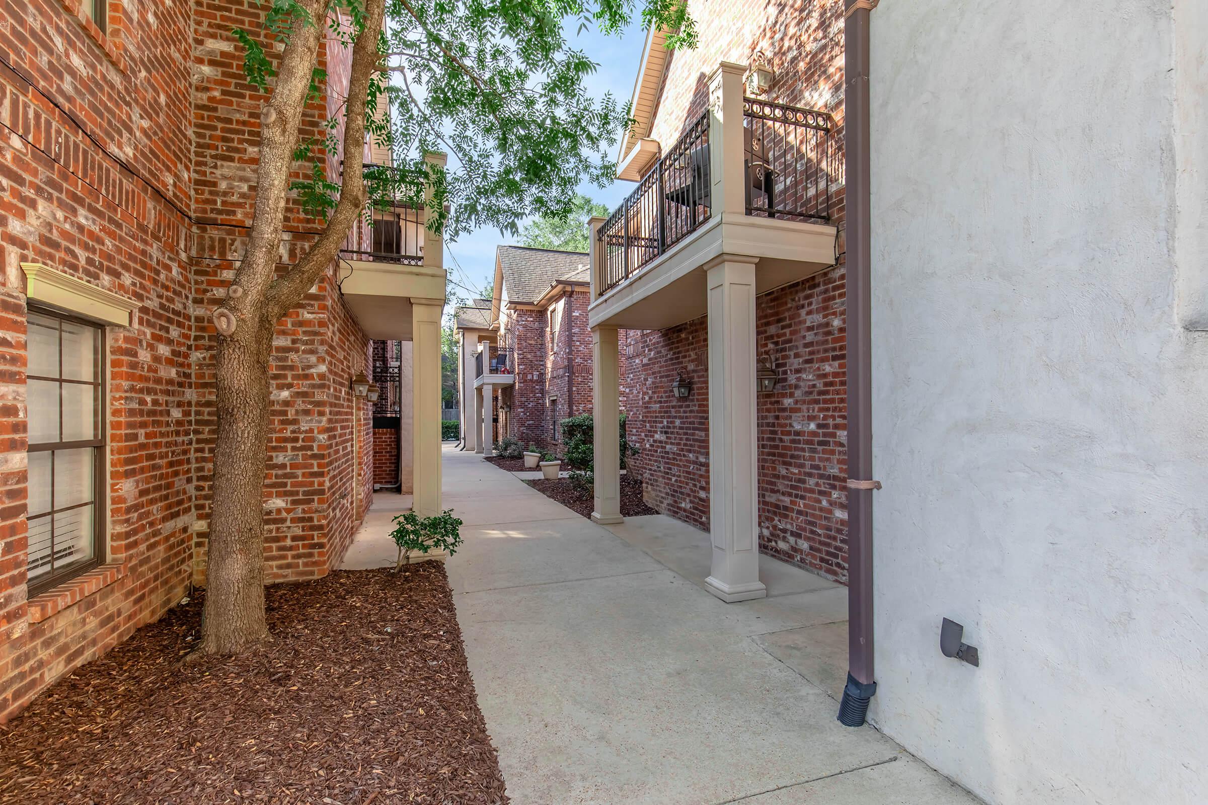 A walkway between brick apartment buildings, featuring trees and well-maintained landscaping. Balconies are visible on some units, and the pathway is lined with mulch. The scene is bright and inviting, suggesting a peaceful residential environment.