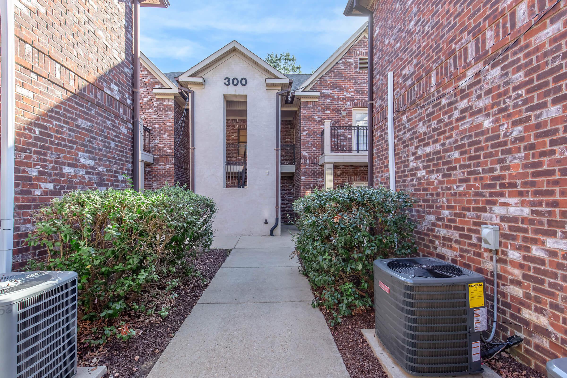 Pathway leading through a brick apartment complex, with green hedges on either side. An air conditioning unit sits to the right, and the building is marked with the number 300. The sky is clear and blue, creating a bright atmosphere in the courtyard-like area.