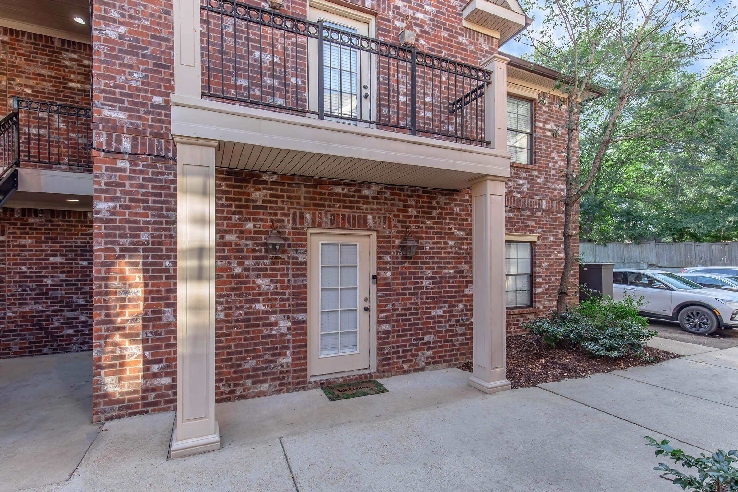 Exterior view of a residential building featuring a brick façade. The image shows a ground-level entrance with a white door and decorative light fixtures on either side. There are balconies above the entrance, surrounded by black wrought iron railings. A small garden area is visible next to the path, with parking spaces in the background.