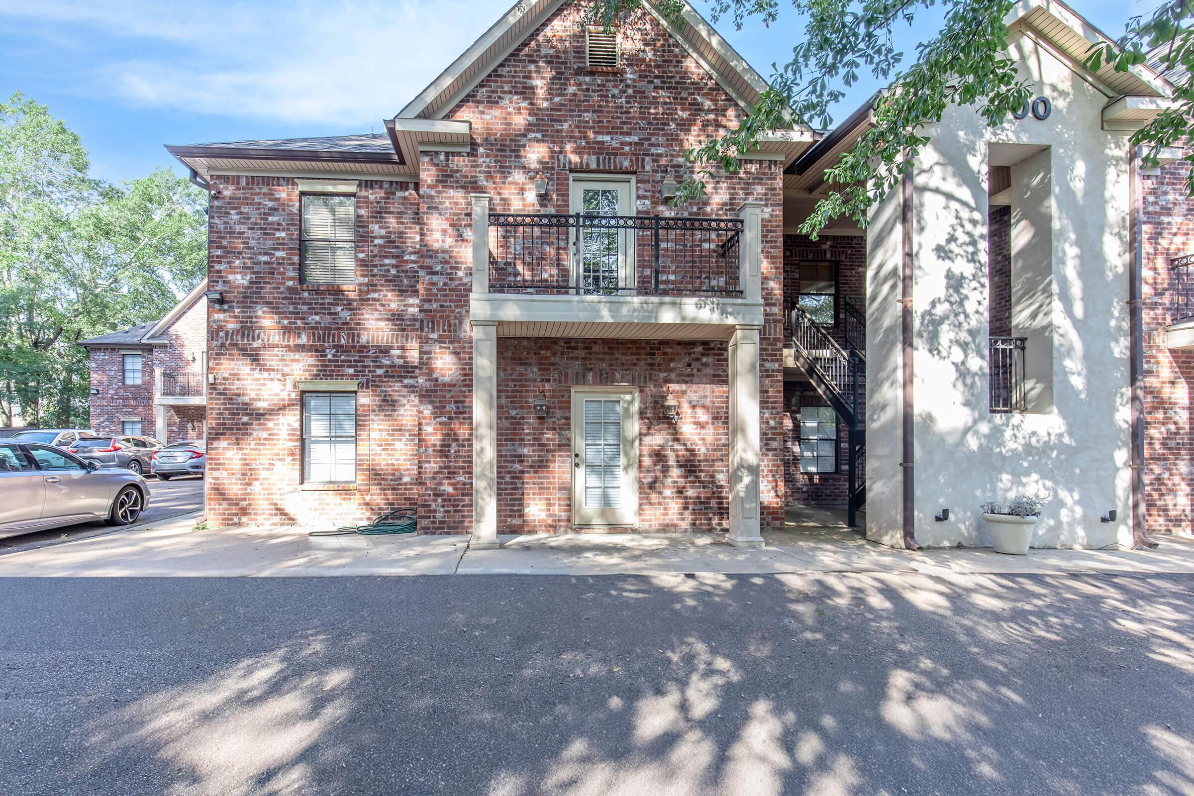 A two-story brick building with a white exterior accent. It features a balcony on the second floor, a front entrance, and outdoor stairs on the side. The area is partially shaded by trees, and there are parked cars in the background. The ground is paved, showing a clear, sunny day.