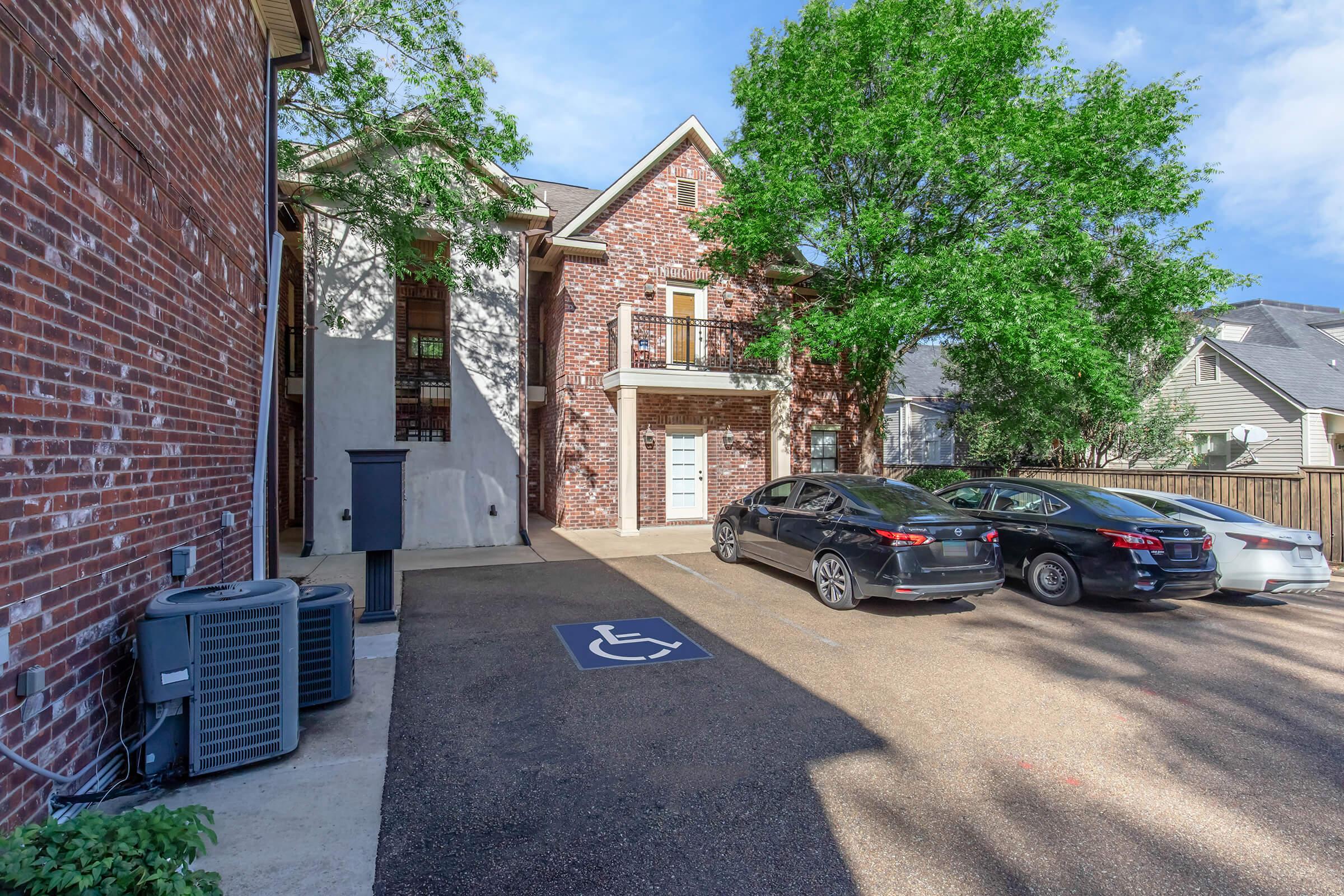 A view of a residential area featuring a brick building with a front entrance. In the foreground, there are two parked cars and a designated handicap parking space. Lush greenery surrounds the area, with trees providing shade. The setting appears well-maintained and inviting.