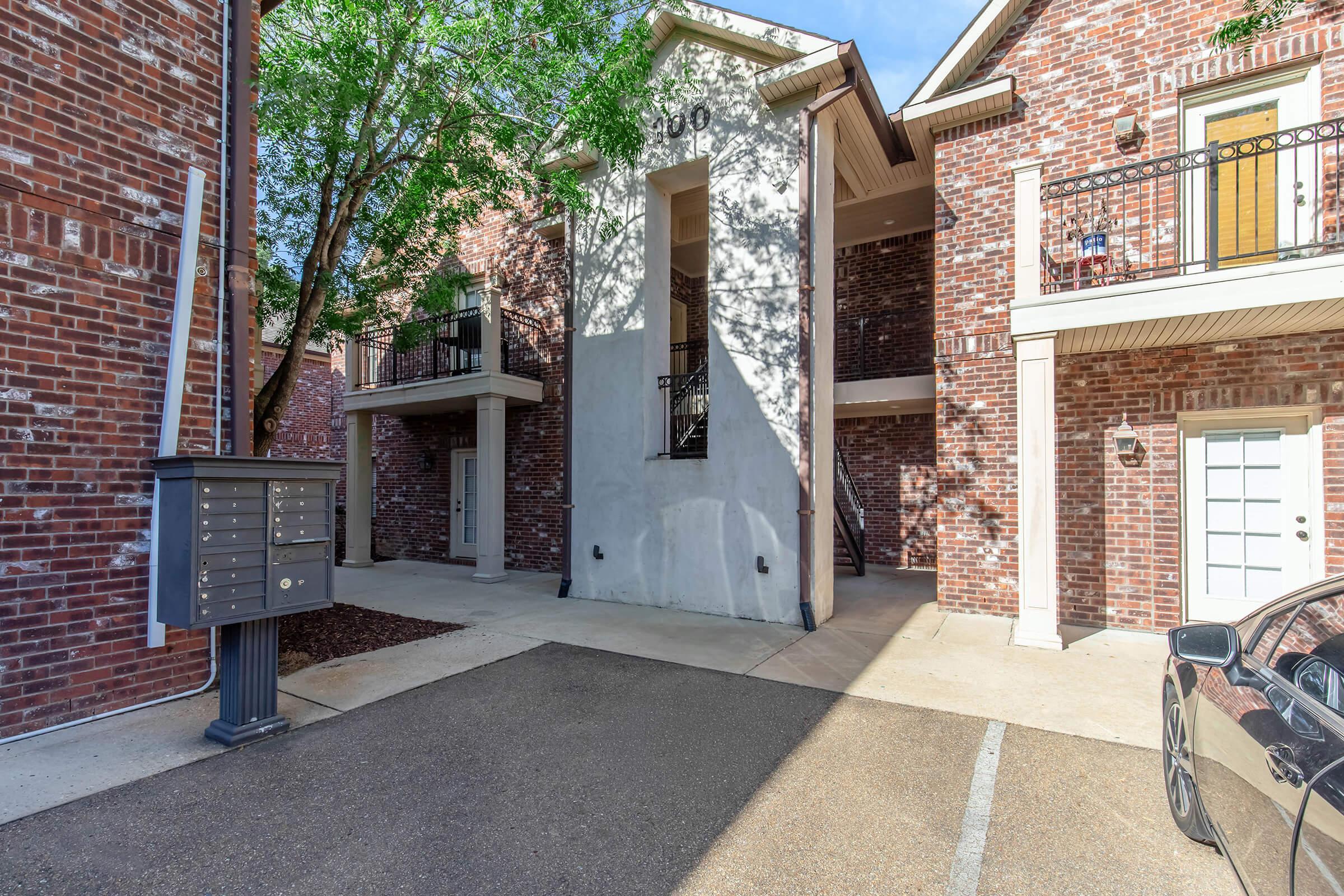 A view of an apartment complex featuring brick exteriors and a landscaping of trees. In the foreground, there is a mailbox stand. The entrance to an apartment unit is visible, framed by two balconies above. The setting is well-lit with clear skies.