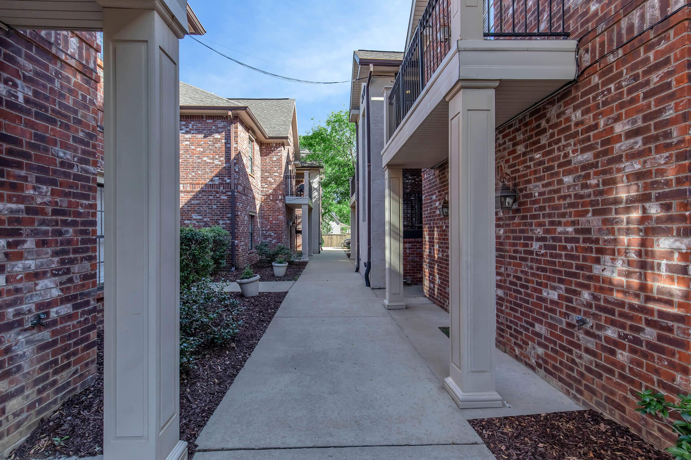 A narrow walkway flanked by brick buildings with columns, featuring landscaped areas with shrubs and potted plants. The sky is clear, and the scene conveys a tranquil, well-maintained residential environment.