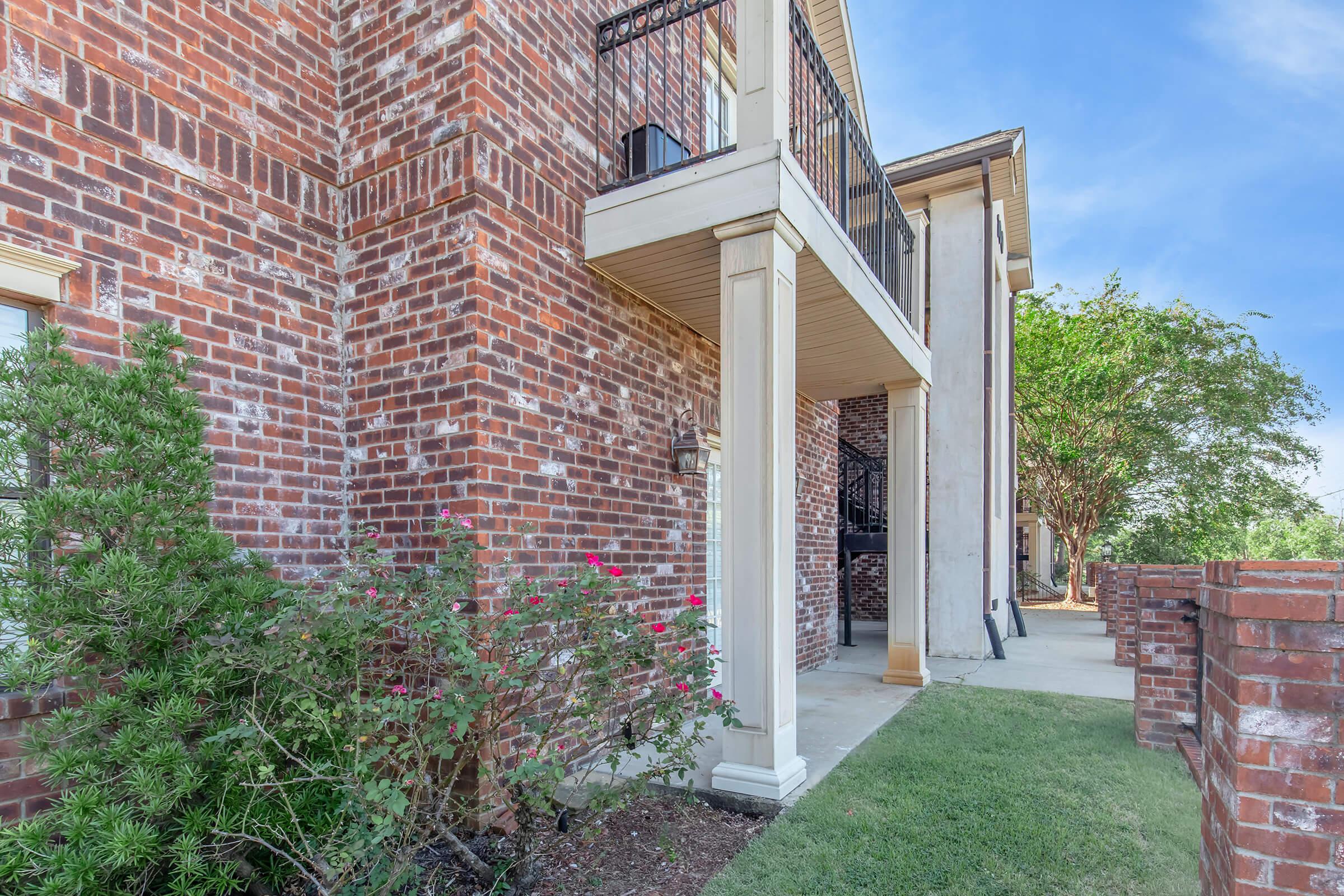 A portion of a brick building featuring a balcony above a covered entrance. The area is landscaped with green grass and flowering bushes, providing a welcoming exterior. A clear blue sky is visible in the background, indicating a bright day.