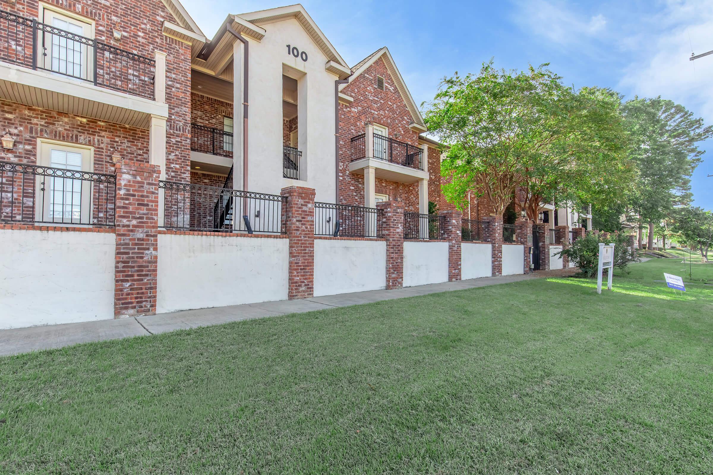 Exterior view of a brick apartment building with multiple balconies, situated on a grassy lawn. The building has a modern design and is lined with black iron railings. A “For Rent” sign is visible in the foreground, indicating available units. The sky is clear with a few clouds.
