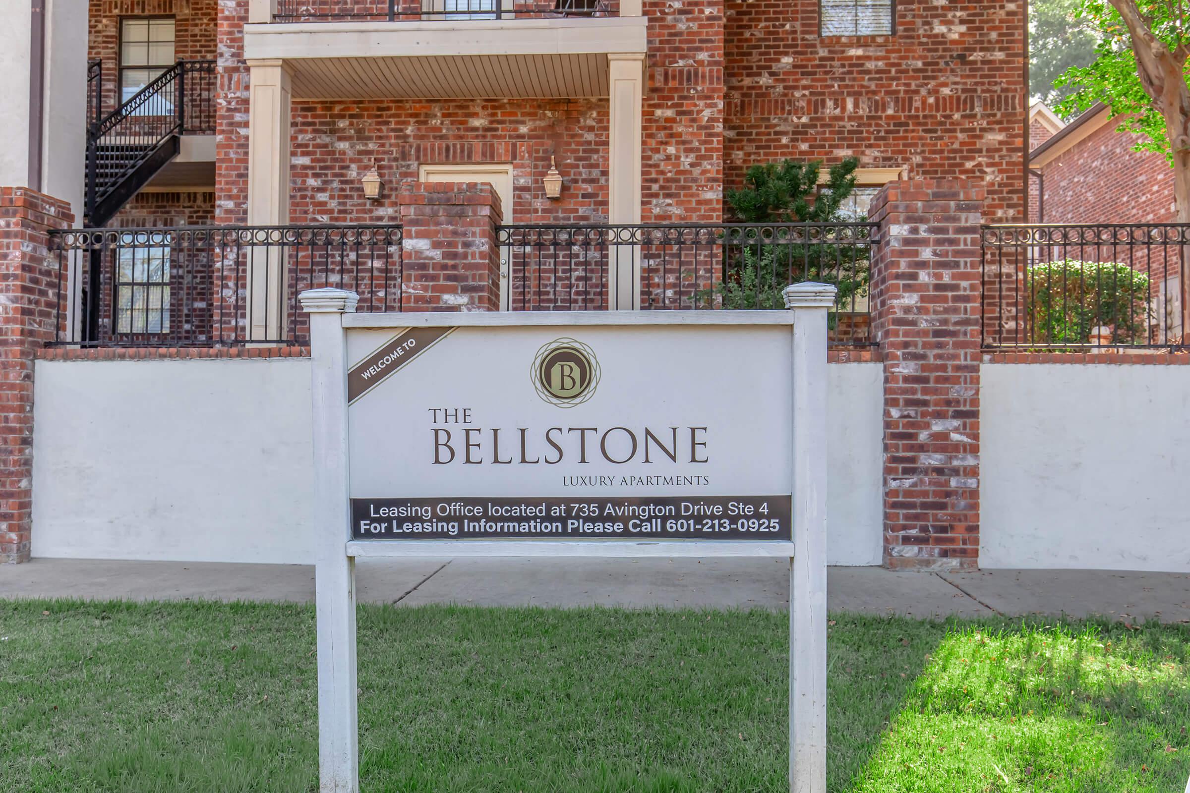 Sign for The Bellstone luxury apartments with leasing office information, located at 736 Avington Drive, Suite 4. Phone number for leasing information: 601-213-0925. The background features a brick wall and greenery.