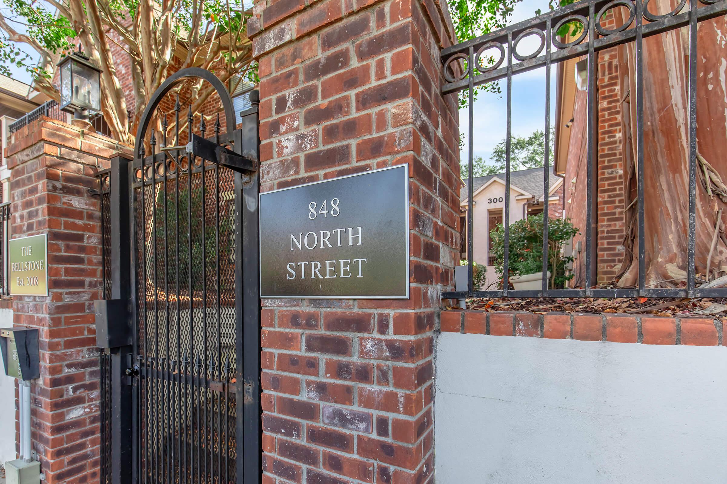 A black sign on a brick wall displaying the address "848 NORTH STREET." In the background, there is a gated entryway with trees and a glimpse of a building. The setting appears to be a residential area with well-maintained landscaping.