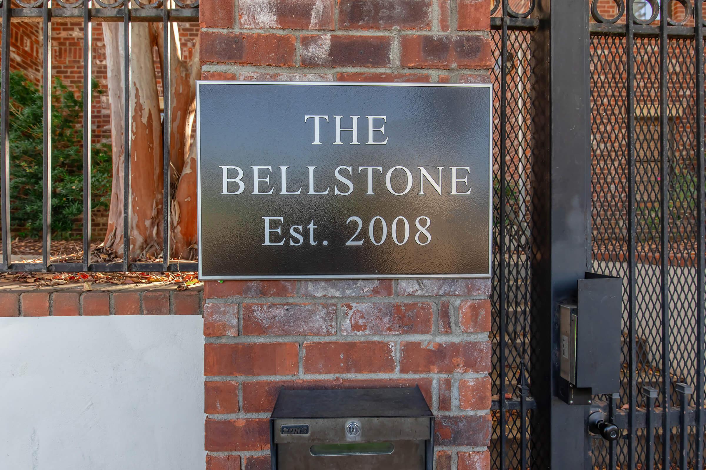 A rectangular sign mounted on a brick wall reads "THE BELLSTONE" in large letters, with "Est. 2008" below it in smaller font. The sign is positioned near a gated entrance, indicating a residential or commercial property. A mailbox is visible at the base of the wall.