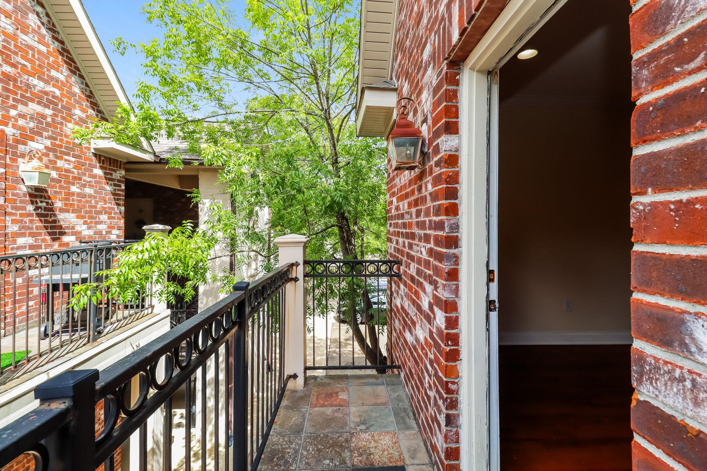 A view from a doorway leading to a small balcony, featuring red brick walls and a decorative railing. There are green trees visible in the background and natural light illuminating the space. The flooring consists of stone tiles, adding to the outdoor appeal.
